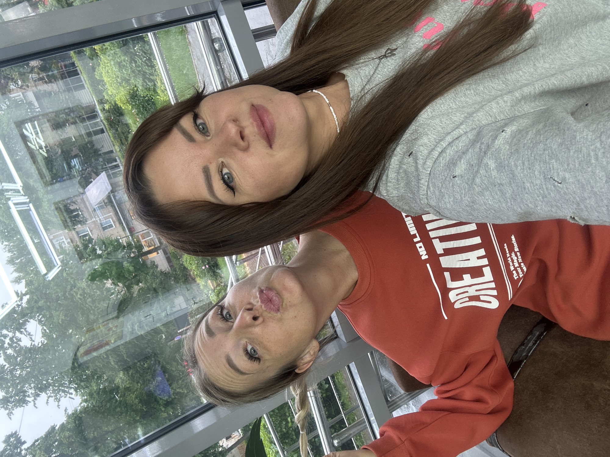 In Chaikovsky, Russia, two women are posing for a selfie indoors, near a large window. The woman on the left, with blonde hair styled into a braid, wears a red sweatshirt with the word "CREATIVE" printed on it. She is puckering her lips towards the camera. The woman on the right, with long brown hair, wears a grey sweatshirt. She is looking directly at the camera with a gentle smile. Through the window behind them, the facade of a brick building and lush green trees are visible, suggesting a somewhat overcast day. The overall scene is casual and friendly.