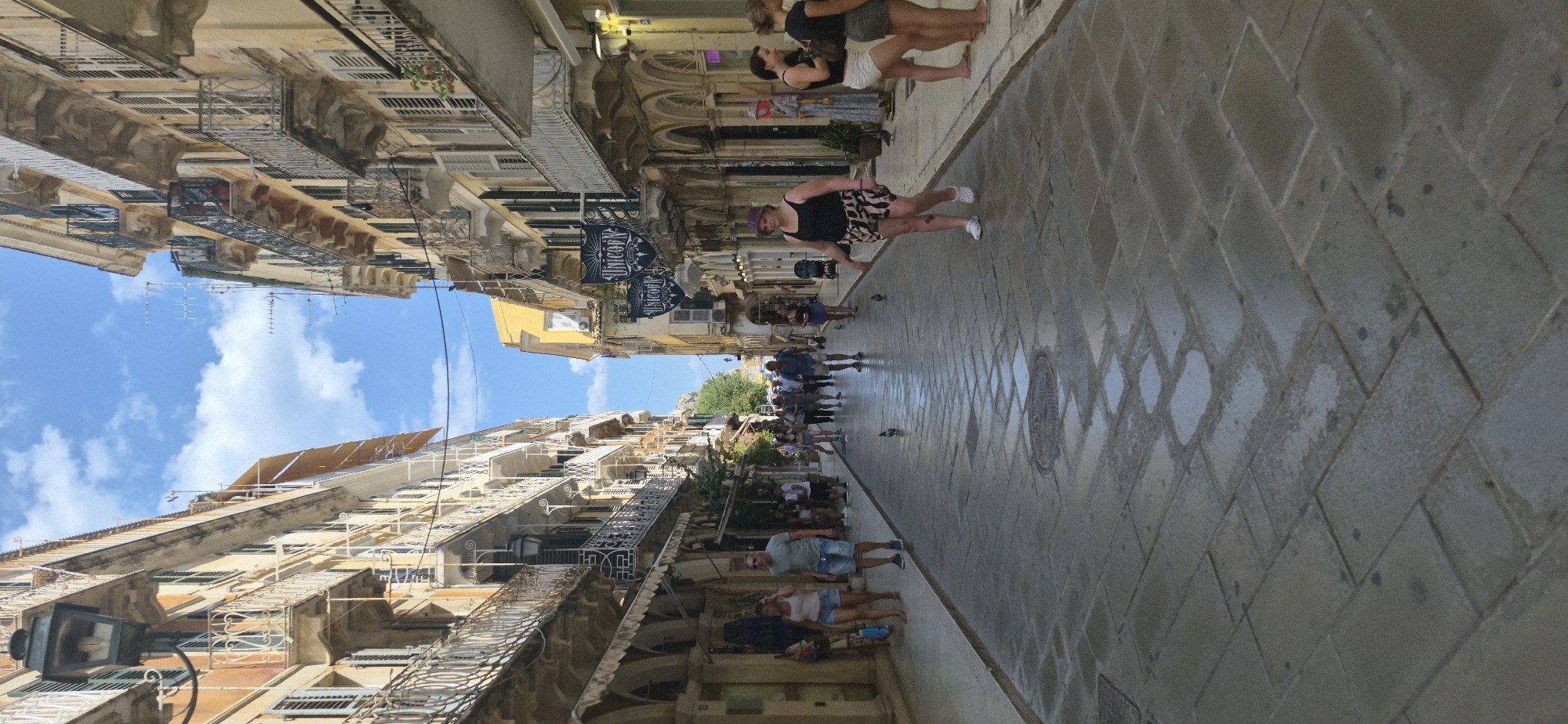 A street scene in Corfu, Greece, captures a perspective looking down a narrow cobblestone street lined with multi-story buildings. The buildings exhibit classical architecture with balconies and shutters, painted in shades of yellow and cream. The sky is blue with scattered white clouds.
Several people are walking along the street. In the foreground, a woman wearing a black tank top and patterned shorts is walking towards the camera. To her right, two women are standing, one in a white skirt and the other in a grey skirt. Further down the street, a group of people are walking in the opposite direction. Some are wearing casual summer clothing.
The street is paved with stone slabs, and parts of it appear wet, reflecting the light. There are a few pigeons walking on the wet cobblestones. Shops are visible along the street, indicated by awnings and signage. One sign reads "UNICORN." The overall atmosphere suggests a sunny day in a tourist area.