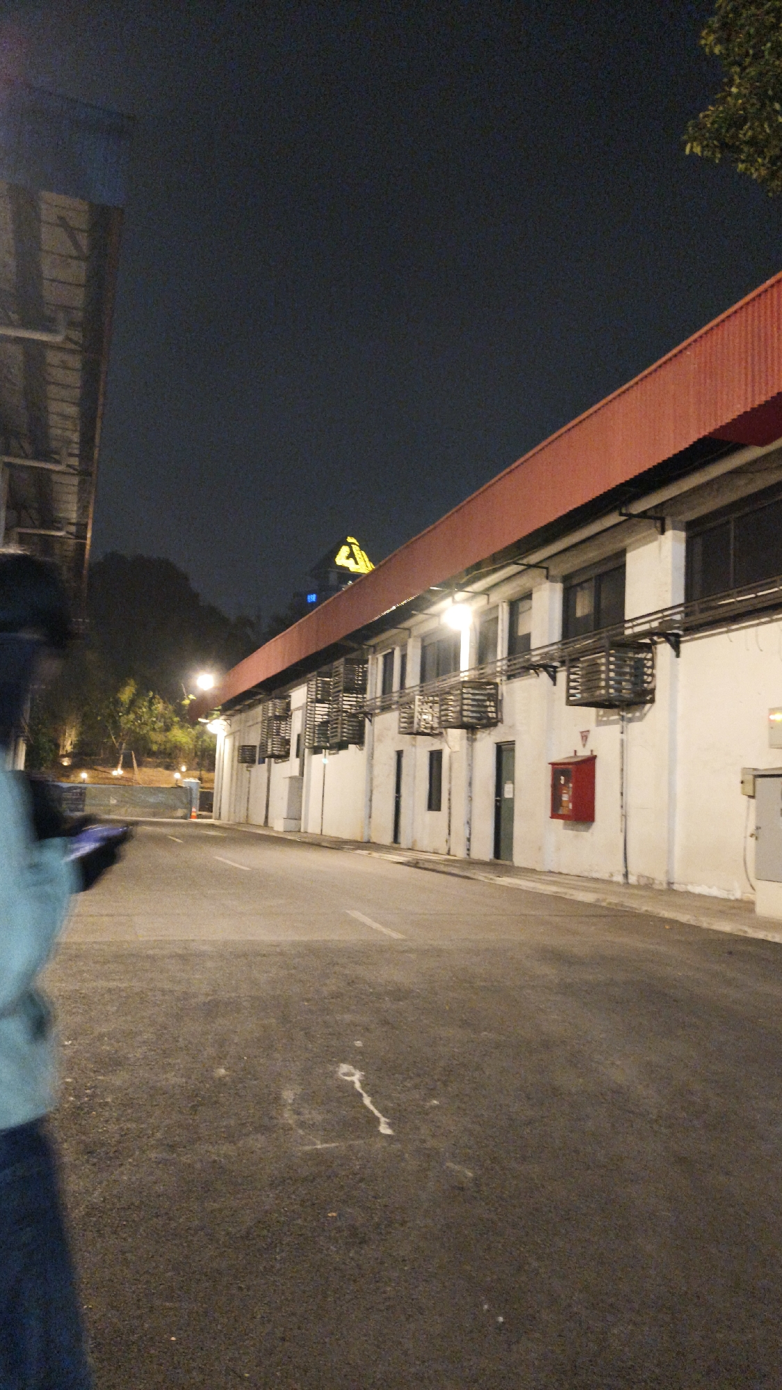 The image depicts a nighttime scene of a long, white, two-story building with a red corrugated metal roof. The building appears to be an industrial or commercial structure, with multiple windows and doorways visible along the ground floor. Several air conditioning units are mounted on the exterior wall. A red fire extinguisher cabinet is also mounted on the wall. The building is situated next to a paved road or parking lot, with white lane markings. In the background, trees and some illuminated streetlights can be seen. A person, partially visible in the foreground on the left, is holding a mobile phone. The overall atmosphere is dimly lit, characteristic of an urban environment at night. The context of "South Jakarta, Indonesia" suggests this may be an industrial or commercial area within that region.