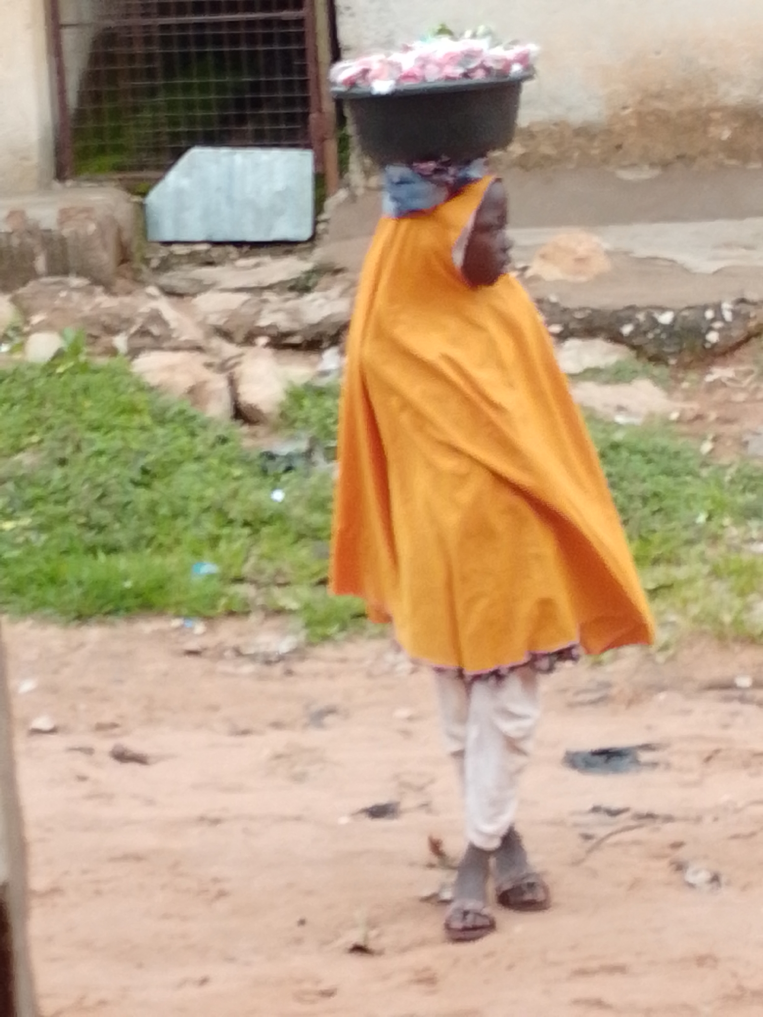 A child wearing an orange head covering and an orange wrap-around skirt walks on a dirt path. The child's face is partially visible and appears to be looking to the right. The child is carrying a black basin filled with pink and white items on their head. In the background, there is a weathered concrete wall with a metal grate, and a patch of green grass with some debris. The ground is composed of reddish-brown dirt with small stones scattered throughout. This scene is located in the city of Tempure, Nigeria.