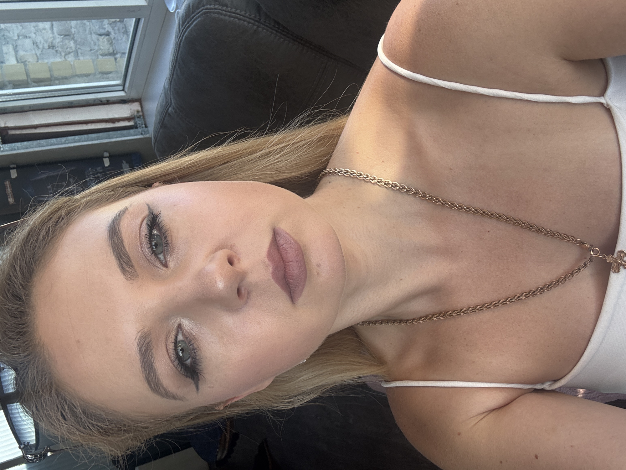 A young woman with blonde hair is pictured in a close-up, head and shoulders shot. She is wearing a white tank top and a gold chain necklace with a pendant. Her makeup includes winged eyeliner and a nude lipstick. The background is blurred, but appears to be an indoor setting with a window showing a brick building.