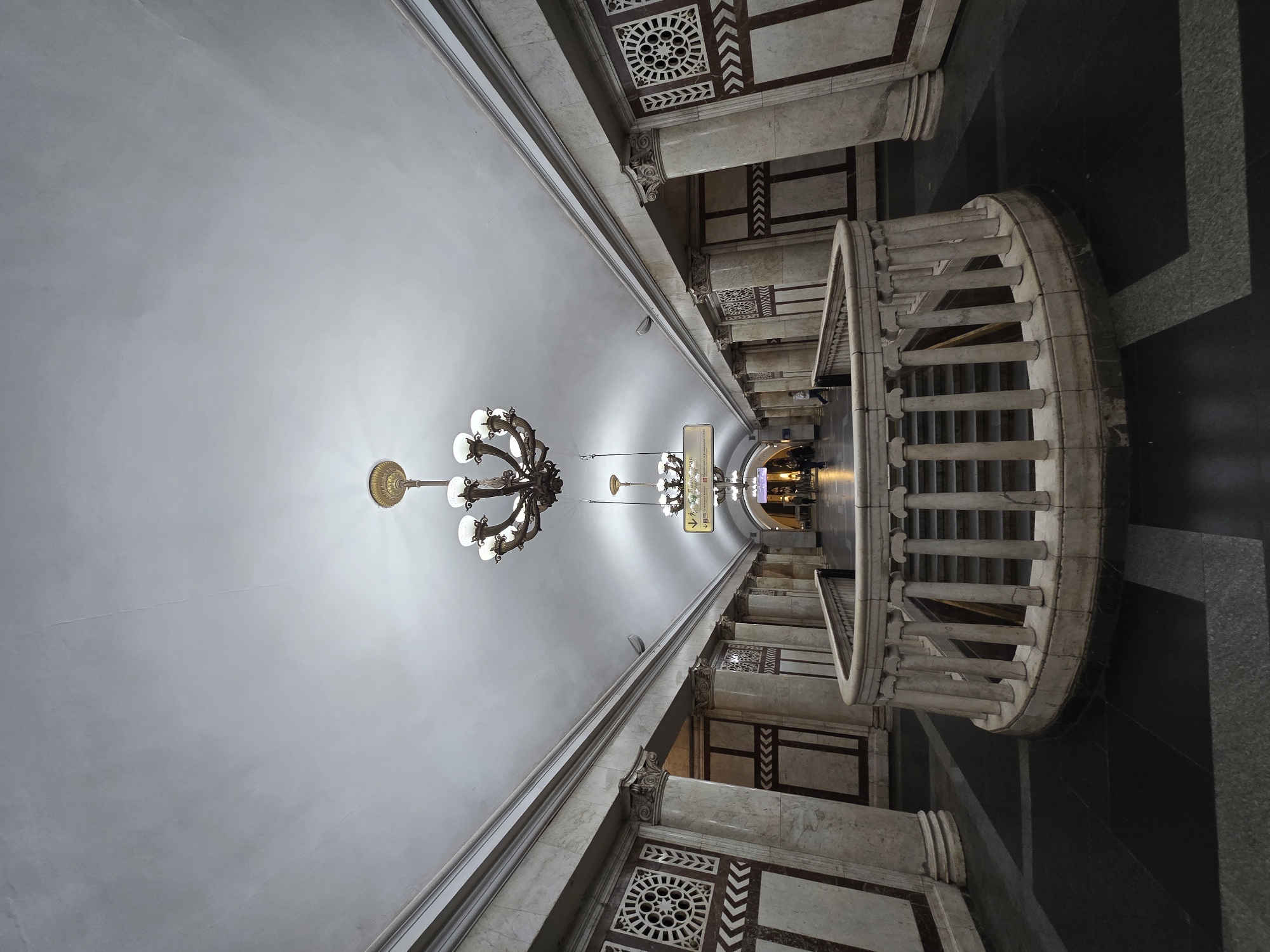 The image shows the Komsomolskaya-Koltsevaya metro station in Moscow, Russia. The perspective is from a low angle, looking up towards the vaulted ceiling. Ornate chandeliers are suspended from the ceiling. The walls are decorated with marble panels, columns, and decorative geometric patterns. A curved white stone staircase with balusters is visible on the right. The floor is patterned with dark and light gray tiles. The station appears mostly empty, with a few blurry figures visible in the distance near the end of the platform.