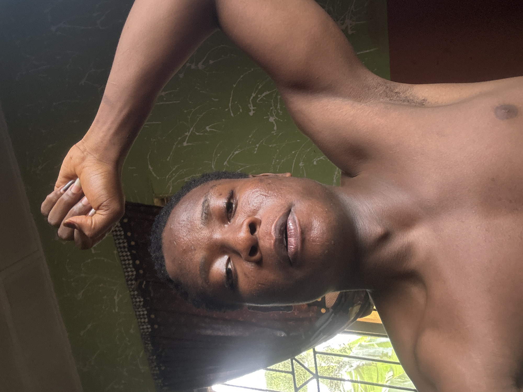 This close-up, low-angle photograph captures a bare-chested person, likely male, with a dark skin tone, looking directly into the camera. The subject's face is central, oriented horizontally in the frame, with a neutral or calm expression. There is a noticeable sheen on their forehead, nose, and lips, possibly indicating sweat or oil. Dark, short hair frames the face, and the lips are slightly parted. One arm is bent and raised, with the hand partially visible at the upper left, while a portion of the chest and shoulder is visible on the right side of the frame.
The background features a distinctive green wall with irregular white splatters or streaks, resembling a decorative paint finish. Below the subject's head, a window is visible, through which bright natural daylight streams in, illuminating lush green foliage outside, suggesting a daytime setting. Part of a patterned, possibly brown and white, fabric or curtain can be seen in the lower background, behind the subject's head. The overall impression is an intimate portrait taken indoors with natural light. There is no visible text.