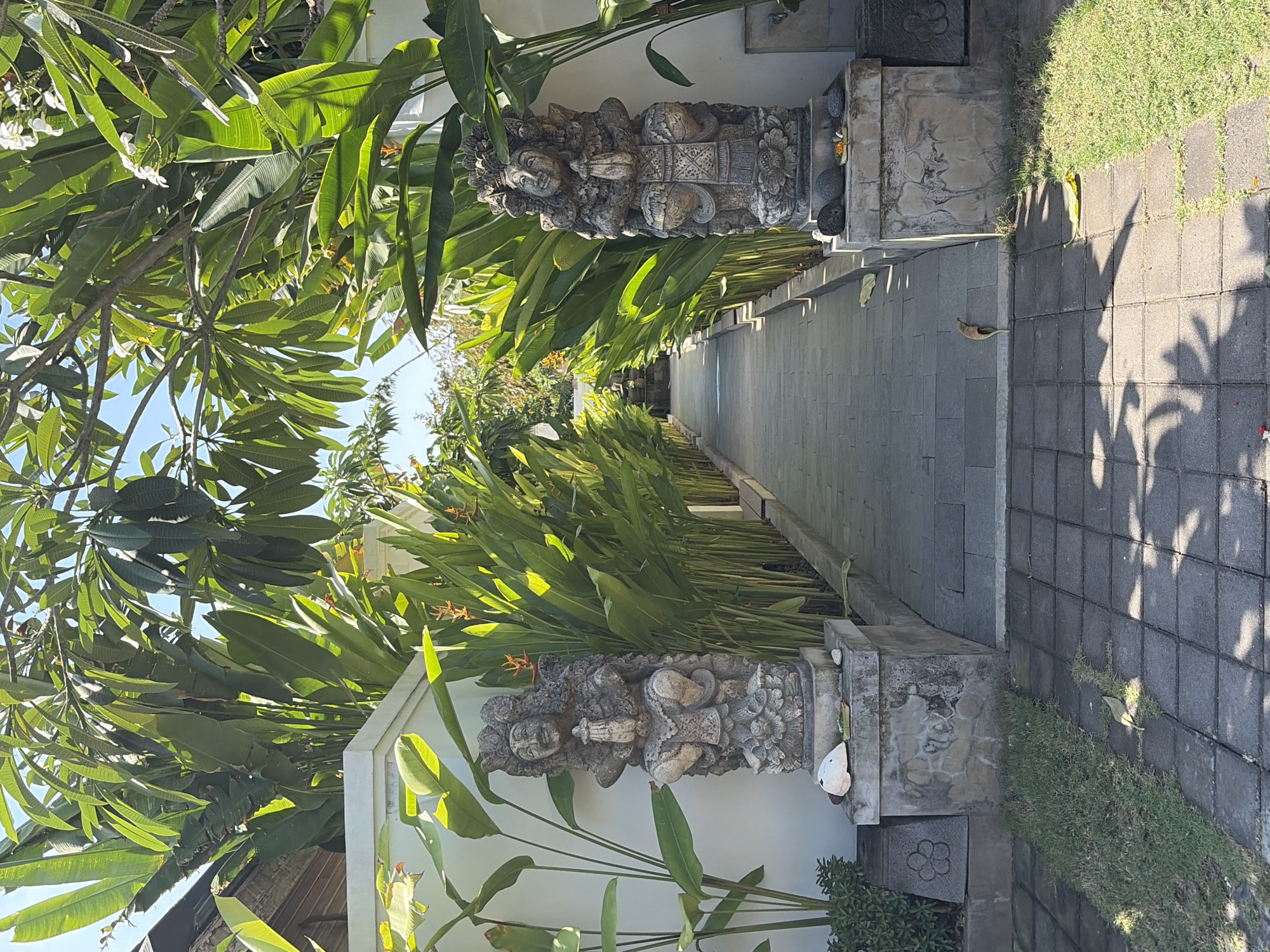 The image depicts a pathway leading into a lush, tropical garden. Flanking the pathway are two intricately carved stone statues, each in a seated, prayer-like pose. These statues are adorned with detailed headdresses and traditional Balinese clothing. Tall, broad-leafed plants, such as Heliconia, line the sides of the pathway, creating a vibrant green corridor. In the background, the pathway recedes into the distance, hinting at more garden elements or a building. The ground is paved with dark, square tiles, and sections of manicured green grass are visible. The scene is bathed in bright sunlight, casting dappled shadows on the path. The overall atmosphere suggests a serene and well-maintained garden, likely part of a resort or private residence in Kuta Selatan, Indonesia, a region known for its beautiful beaches and tropical landscapes.