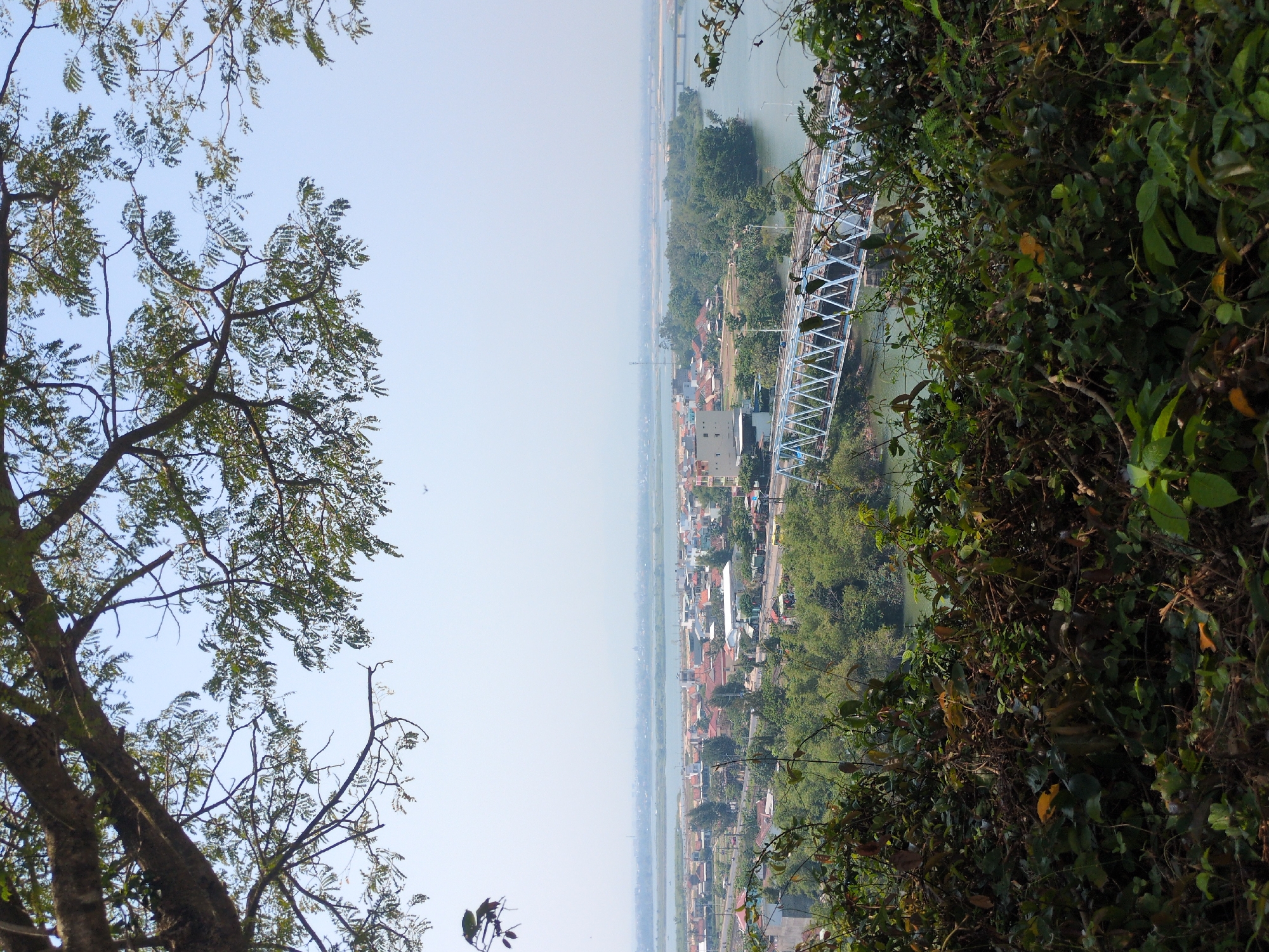 The image depicts a landscape view from an elevated position, possibly a hillside, overlooking an area that appears to be the city of Tuy Hòa, Vietnam. The perspective is obscured by vegetation on both sides, with a tree on the left and a dense collection of leaves and branches on the right. A river runs through the center of the frame, bisecting the city. A blue truss bridge spans the river, connecting sections of the town. Further in the distance, another bridge is visible. Buildings and houses are spread along the landscape on both sides of the river. The sky is clear and hazy, suggesting a bright day.