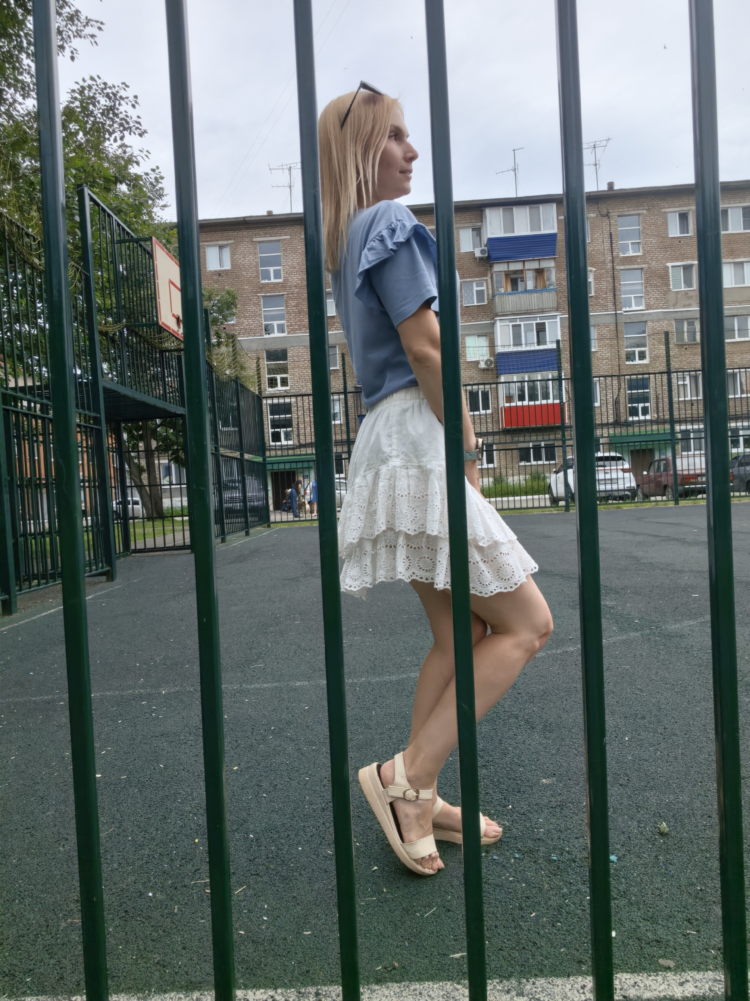 The image shows a woman standing on an outdoor sports court in a city. She is wearing a blue t-shirt and a white ruffled skirt, with beige sandals. She is partially obscured by green metal bars, which appear to be part of a fence surrounding the court. In the background, there is a brick apartment building, a basketball hoop, and some trees. The sky is overcast. The location is the courtyard of an apartment complex in Sterlitamak, Russia.