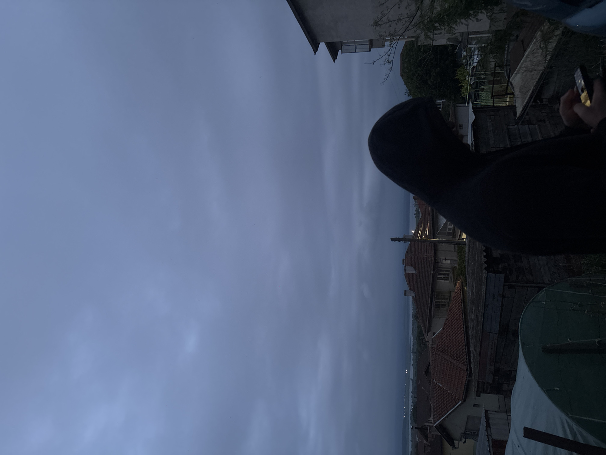 The photograph depicts a hooded figure holding a mobile phone, likely taking a picture or video. In the background, there is a cityscape under a cloudy sky, featuring buildings with red-tiled roofs, a body of water, and distant lights. The scene is potentially located in Burgas, Bulgaria.