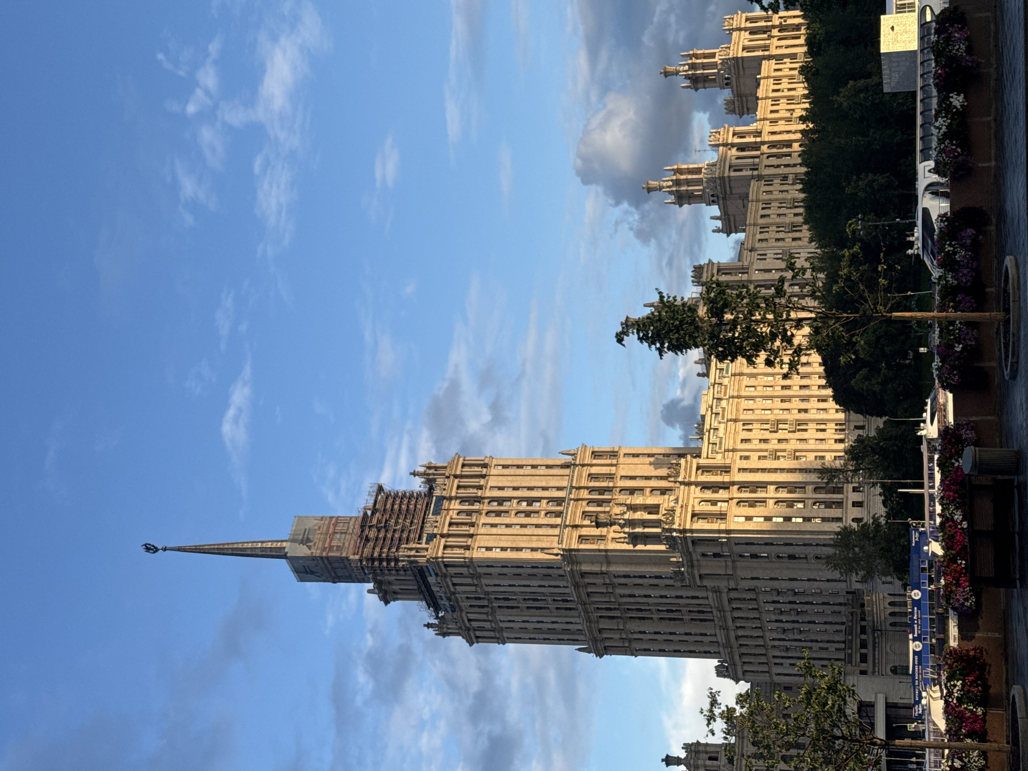 The image shows a sunlit view of the Ministry of Foreign Affairs building in Moscow, Russia, under a partly cloudy blue sky. The iconic skyscraper, a stalinist-era architectural masterpiece, dominates the frame with its intricate facade and tall spire. The lower portions of the building appear to be undergoing some renovations, with scaffolding visible around the upper levels of the main tower.
In the foreground, a line of trees and flower beds add a touch of greenery and color to the scene. A modern boat with a streamlined design is docked along the waterfront, with its windows reflecting the sky. Several other buildings, also in a similar architectural style, extend to the right of the main structure, creating a sprawling urban landscape. The lighting suggests it is either early morning or late afternoon, casting a warm glow on the buildings. The overall impression is one of grandeur and historical significance, characteristic of Moscow's prominent landmarks.