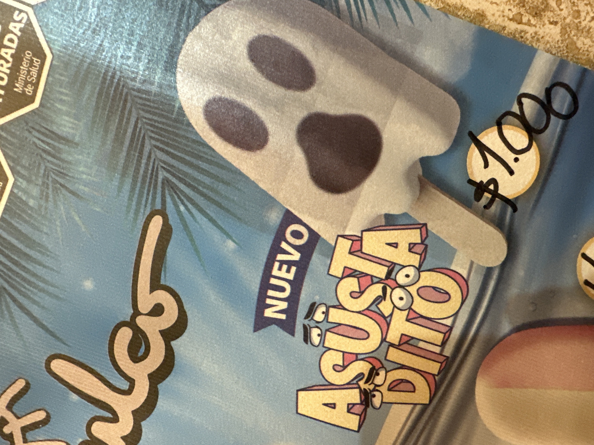 The image shows a promotional advertisement for a "Nuevo Asusta Dito" popsicle, featuring a cartoon ghost-shaped ice cream. The price of the popsicle is indicated as $1.000. The advertisement also displays the phrase "Ministerio de Salud" partially visible. The location is Villa Adelina, Argentina.