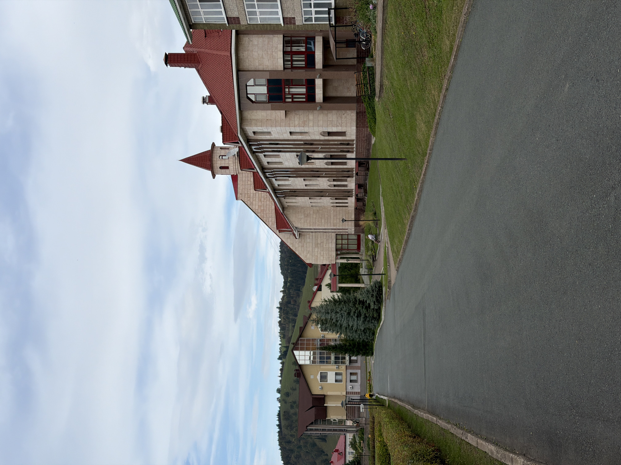 A paved road leads through a residential area with multi-story buildings on the right side of the frame. The buildings have red-tiled roofs and light-colored stone exteriors. Some buildings feature tall windows and architectural details like turrets. Lush green grass covers the slopes alongside the road and around the buildings. Several evergreen trees are planted near the buildings. In the background, rolling hills covered in trees are visible under a cloudy sky. A few bicycles are parked near one of the buildings. One person is seated in a wheelchair on a paved path near the buildings.