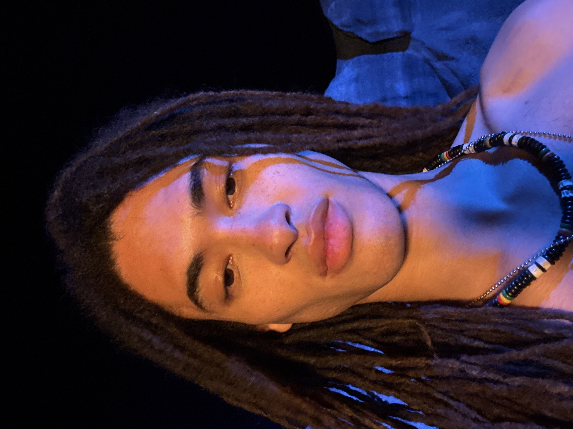 A person with dark brown dreadlocks, light brown skin, and a beaded necklace is the primary subject in this portrait-style image. The person is lying down, facing the camera with a neutral expression. The lighting is uneven, with orange illumination on the face and blue lighting towards the background. A dark shape resembling rocks can be seen behind the person. There is no information in the image to determine the location is Teguise, Spain.