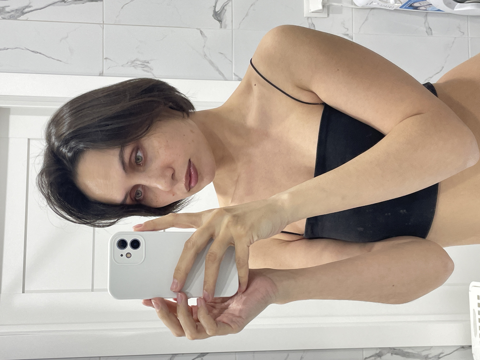 A person with short, dark hair and fair skin is holding a white smartphone and taking a selfie in a bathroom. The person is wearing a black strapless top with thin straps. The background consists of white tiled walls with gray marble-like veining. A white door frame and a partial view of other bathroom items can be seen. The location cannot be determined from the image alone.