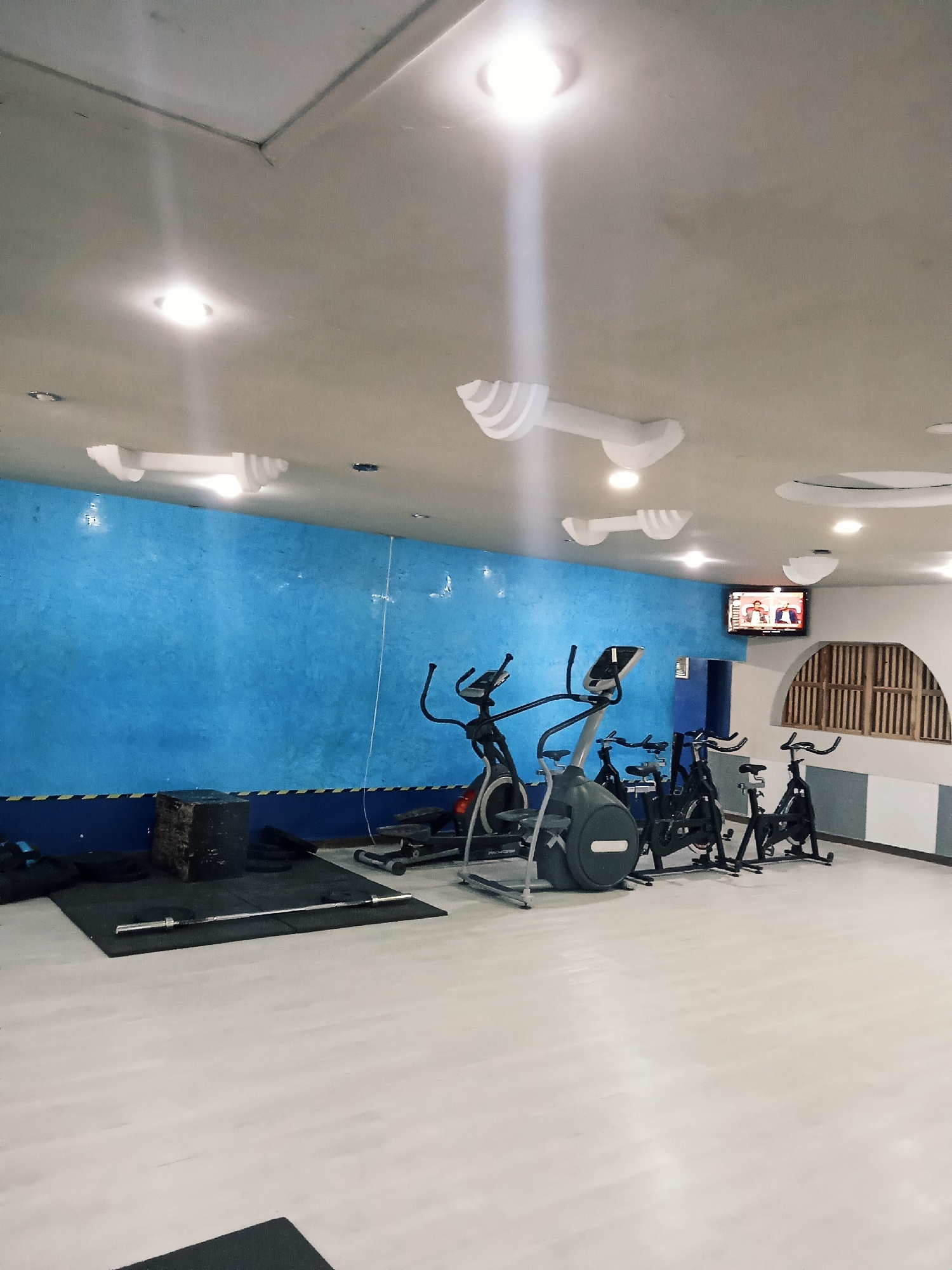 The image depicts a gym located in Altagracia, Venezuela. The gym features a blue accent wall with painted white stars, a gray and white striped wall, and exercise equipment including elliptical machines, stationary bikes, a barbell, and a plyometric box. A television is mounted on the wall, displaying a broadcast. The gym has a white floor and a white ceiling with unique decorative light fixtures. No people are visible.