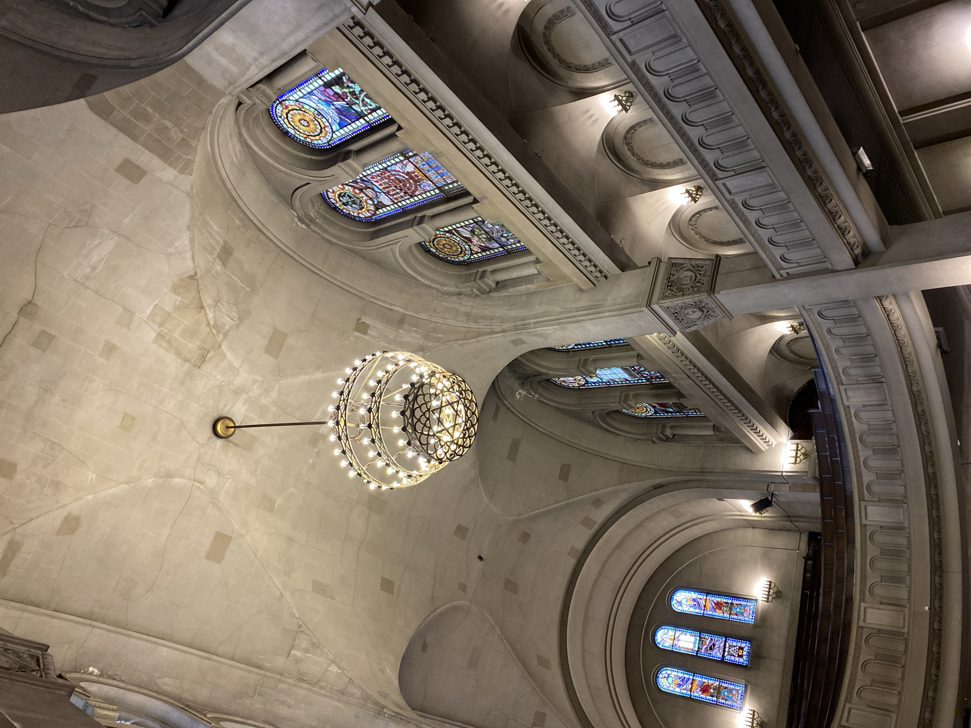 The image depicts the interior of a building with arched ceilings and stained glass windows. A large, spherical chandelier hangs from the center of the ceiling. The walls are constructed of light-colored stone. Ornate molding and decorative features are present. The scene is likely the interior of a church or cathedral. The image was probably taken in Buenos Aires, Argentina.