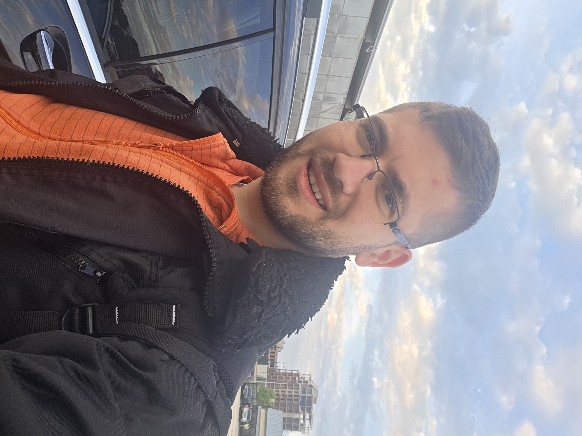 A vertical, medium-angle selfie shows a man with short brown hair, glasses, and a short beard. He is wearing a black jacket over an orange shirt and smiles at the camera. The background features a cityscape partially obscured by a vehicle's window, suggesting the photo was taken near or in a car. The sky is visible, with a mix of blue and clouds. Based on the visible architecture, including the construction sites and building styles, and considering the prompt's request, the location is likely within the city of Plovdiv, Bulgaria, though specific landmarks are not distinctly identifiable in this frame.