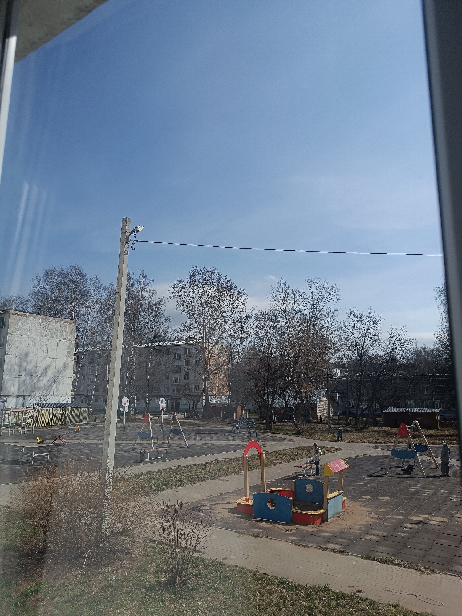 The image depicts a playground in a city, likely in a former Soviet Union country, evidenced by the style of buildings. The sky is clear and blue. Several buildings of typical Soviet architecture are visible in the background. Several bare trees are scattered around the playground.
The playground features a variety of equipment, including swings, climbing frames, and a sandbox structure with bright red, blue, and yellow paint. Two adults stand near the swings, one of them holding a child. A single person stands near the sandbox. A street light is standing on the left of the frame with multiple wires connected to it.