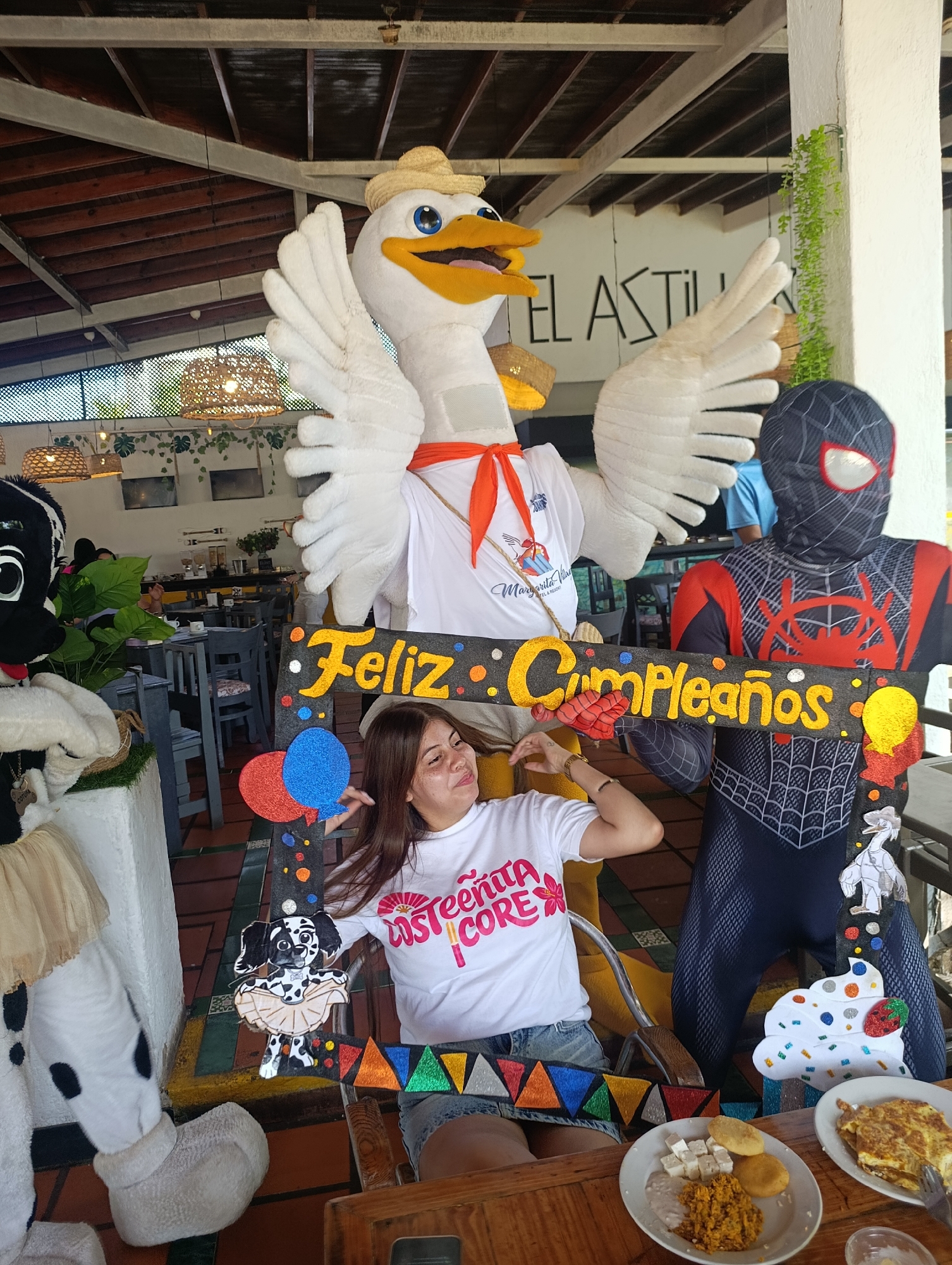 A woman is sitting in a chair posing behind a "Feliz Cumpleaños" (Happy Birthday) sign held by a person in a Spider-Man costume and a large goose mascot wearing a straw hat and an orange scarf. To the left of the woman is a person in a Dalmatian mascot costume. The woman is wearing a white t-shirt with pink and red writing and a pair of denim shorts. A plate of food is on the table in front of her.
The scene appears to be inside a restaurant in Porlamar, Venezuela. In the background are tables and chairs, light fixtures, and plants. A sign reading "el astil" is visible. The logo on the goose mascot's shirt indicates the Margarita Village Hotel & Resort.