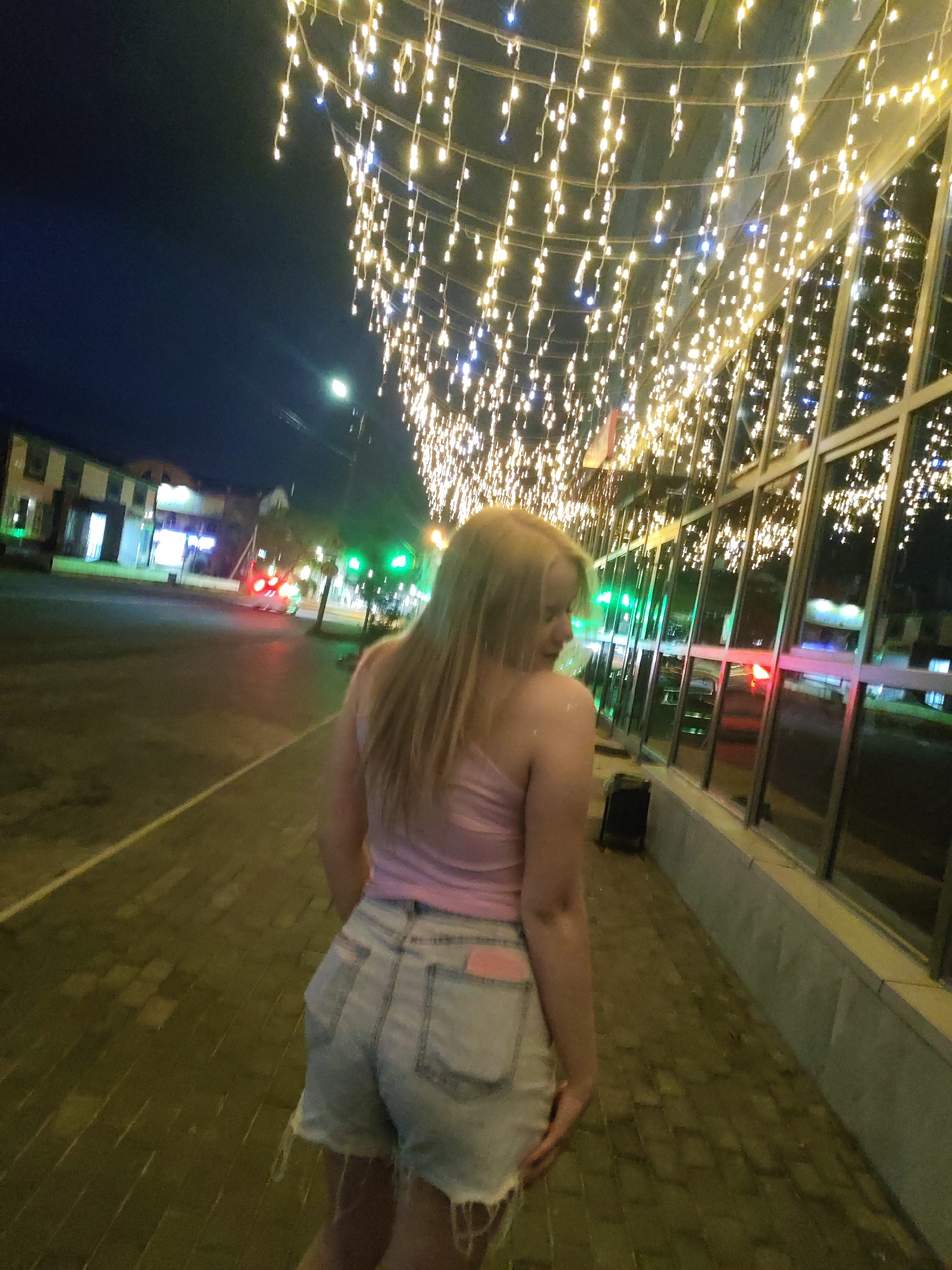 The image depicts a young woman from behind, walking on a paved sidewalk at night. She has blonde hair and is wearing a pink tank top and denim shorts. She is looking over her left shoulder, slightly to the right. The background shows a street with buildings and cars. The scene is illuminated by streetlights and numerous string lights draped overhead, creating a festive or decorative atmosphere. The overall setting appears to be an urban environment.