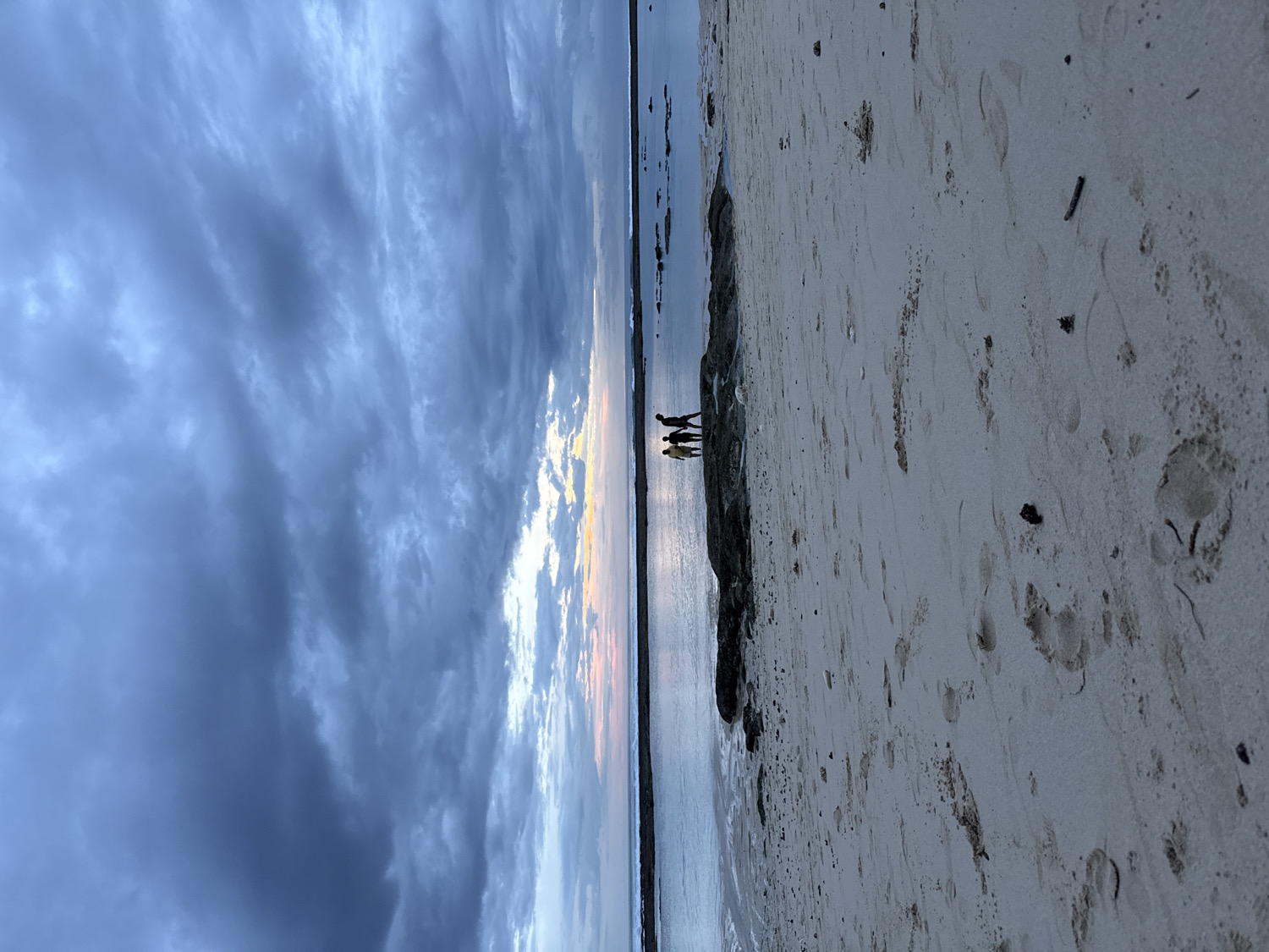 The image captures a dramatic beach scene during either sunrise or sunset in Kuta Selatan, Indonesia. The sky occupies a significant portion of the frame, filled with dense, dark grey and blue clouds, interspersed with brighter patches of orange and yellow light, indicating the position of the sun just below the horizon or breaking through the cloud cover.
In the mid-ground, the calm, reflective waters of the ocean stretch horizontally, meeting the sandy shore. A dark, low-lying rocky outcrop or natural barrier extends along the water's edge, separating the ocean from the expanse of beach.
Three human figures are silhouetted against the bright, reflective line where the water meets the distant sky. They appear to be standing on or near the rocky formation, observing the horizon. Their forms are indistinct due to the backlighting.
The foreground consists of a wide stretch of light-colored sand, marked with numerous footprints of varying sizes and small pieces of natural debris, suggesting recent activity. The overall atmosphere is serene and contemplative, enhanced by the striking sky. There is no visible text in the image.
