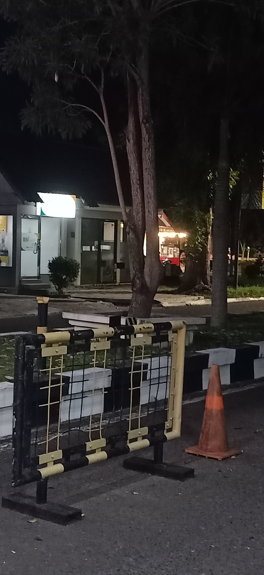 This is a nighttime outdoor photograph of a street scene in Tambontunong, Indonesia. In the foreground, a temporary black metal barrier with yellow accents and a wire mesh is set up on the asphalt. To the right of the barrier, an orange traffic cone stands on a pallet. Behind the barrier, a sidewalk with black and white striped curbs is visible, with some white rectangular objects on it. Further back, a building with a lit sign displaying "BSI" is partially visible on the left. To the right of the building, a food stall with warm yellow lighting is operating. Several trees with dark foliage are scattered throughout the scene, their branches extending over the area. The overall lighting is dim, consistent with nighttime conditions, with artificial lights from the building and food stall providing the primary illumination. No people are clearly discernible in the image.