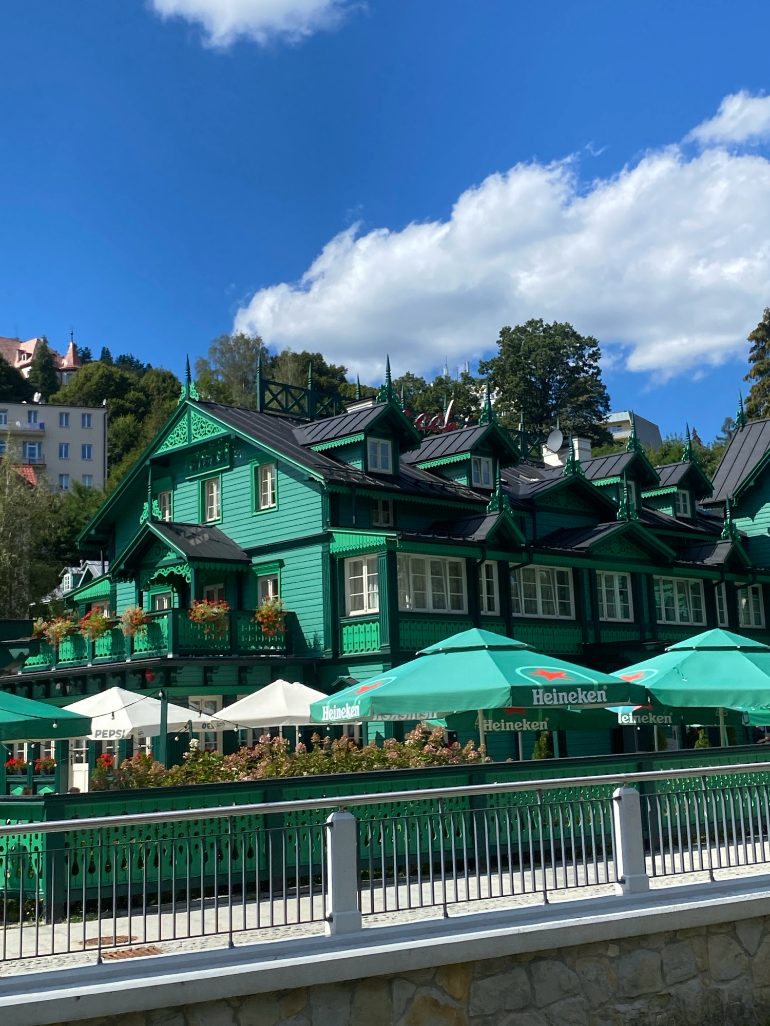 The image shows a brightly colored green building with intricate white trim and detailing, set against a backdrop of trees and a blue sky with white clouds. The building appears to be a restaurant or cafe, with outdoor seating covered by large green umbrellas. The umbrellas are branded with "Heineken" and "Pepsi" logos, indicating advertising for these beverage companies. Flower boxes with red flowers are visible on the balconies of the building. In the background, other buildings and lush green trees climb a hillside, suggesting a scenic, possibly mountainous, location. The foreground features a metal railing and a stone wall. This building is recognizable as the historic "Biały Dom" (White House), a landmark in Krynica-Zdrój, Poland, known for its distinctive architecture and spa town ambiance. There are no people visible in the image.
