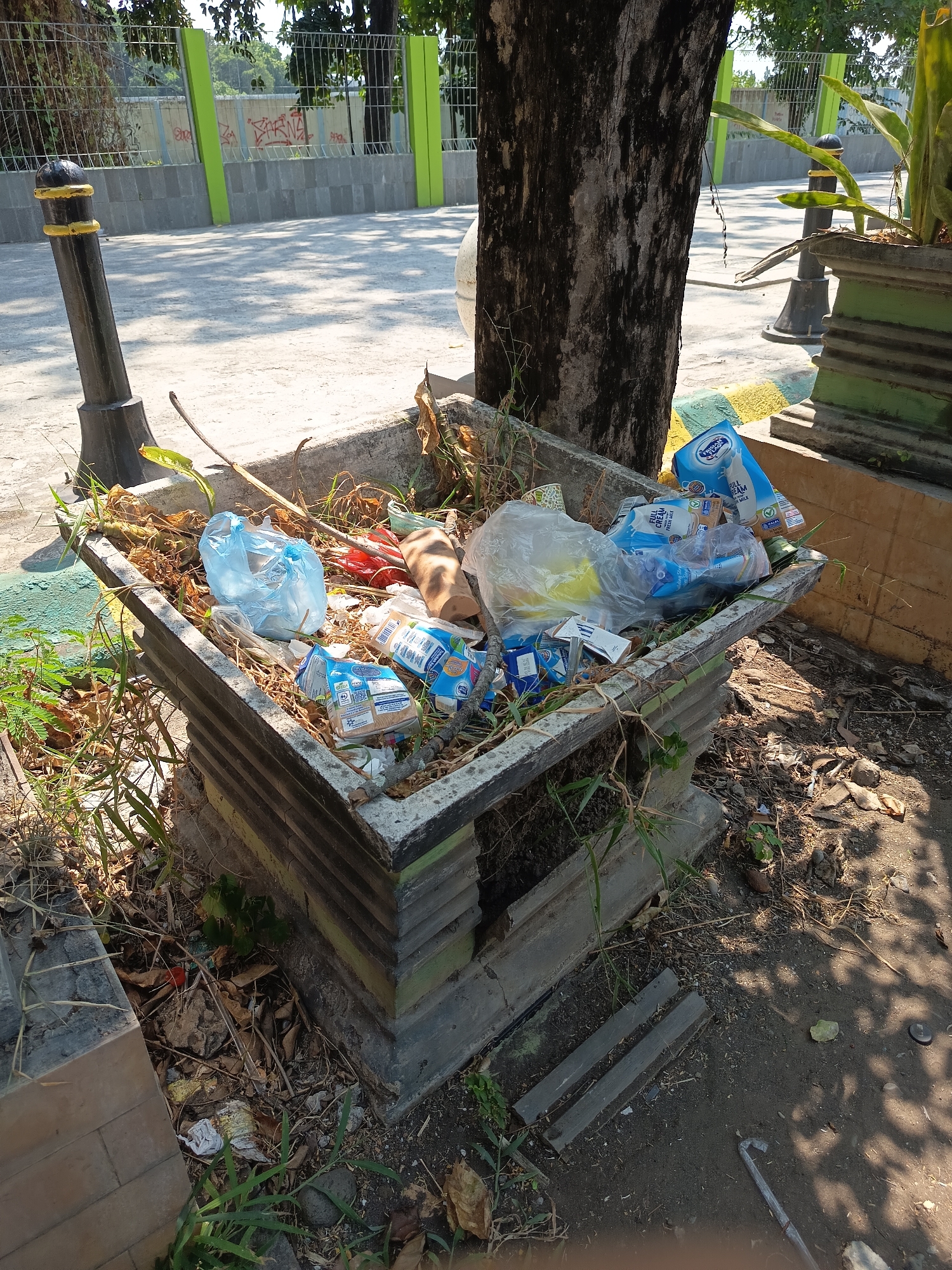 In the city of Kandangjati Kulon, Indonesia, a concrete planter, likely intended for decorative foliage, has been repurposed as a makeshift trash receptacle. The planter, which sits on a weathered, stepped base, is overflowing with discarded items. Among the visible refuse are several blue and white cartons of what appears to be milk, some crumpled plastic bags, dry leaves, twigs, and other unidentifiable debris. The planter itself shows signs of wear, with a rough, textured concrete finish. It is situated outdoors, on a paved area, with a tree trunk visible to the left and a green metal fence with graffiti in the background. The ground surrounding the planter is also littered with dry leaves and dirt.