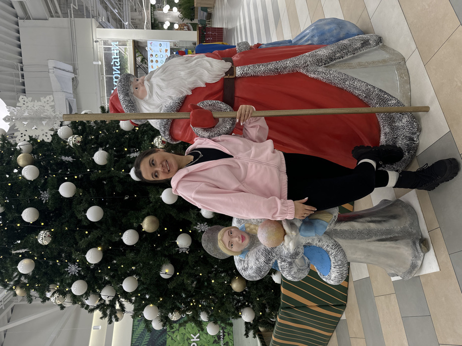 An indoor scene depicts a woman posing amidst holiday decorations within a commercial complex identified by a partial sign as "Okruzhnoy Park" in Yudino, Russia. The woman, with dark hair, stands with her left leg bent, facing slightly left and smiling. She wears a light pink zippered hoodie over a black top, black leggings, white socks, and black platform boots. Her left hand holds a tall staff topped with a snowflake, which is part of a large, red-robed Ded Moroz (Santa Claus) statue positioned to her right. To the woman's left stands a large statue of Snegurochka (Snow Maiden) in a silver and blue costume, holding a golden orb. Behind the woman is a large, artificial Christmas tree adorned with white and gold spherical ornaments and lights, enclosed by a green and gold striped planter box. The environment features a light-colored tiled floor, high ceilings with artificial lighting, and distant commercial signage, including Russian text ("КУРИНЫЙ" - Chicken), suggesting a food court or retail area.