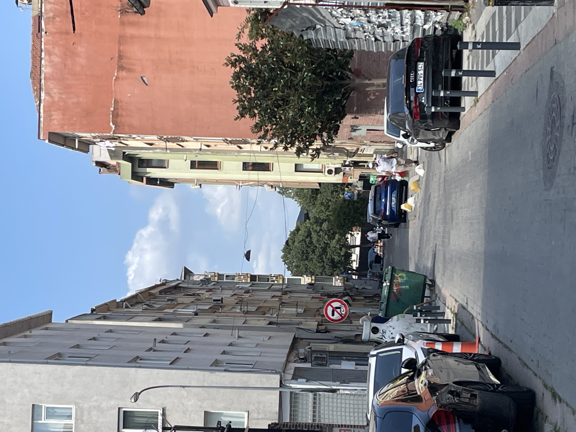 The image shows a narrow street in Kadıköy, Turkey. On the left side of the street are tall, light-colored apartment buildings with numerous windows and balconies. On the right side, there is a large salmon-colored building adjacent to a lighter green building. A large tree with dense foliage stands in front of the salmon-colored building. The street is paved with cobblestones. Several cars are parked along the right side of the street, including a black Audi with license plate "34 PVG 542". A white SUV is parked next to it. A person is walking away from the camera down the street, wearing a light-colored shirt and pants. In the foreground, there are orange traffic cones and trash bins. A red and white circular sign with a diagonal line through it, indicating "no entry," is visible on the left side of the street. The sky is blue with some scattered clouds. The scene appears to be a typical urban environment on a sunny day.