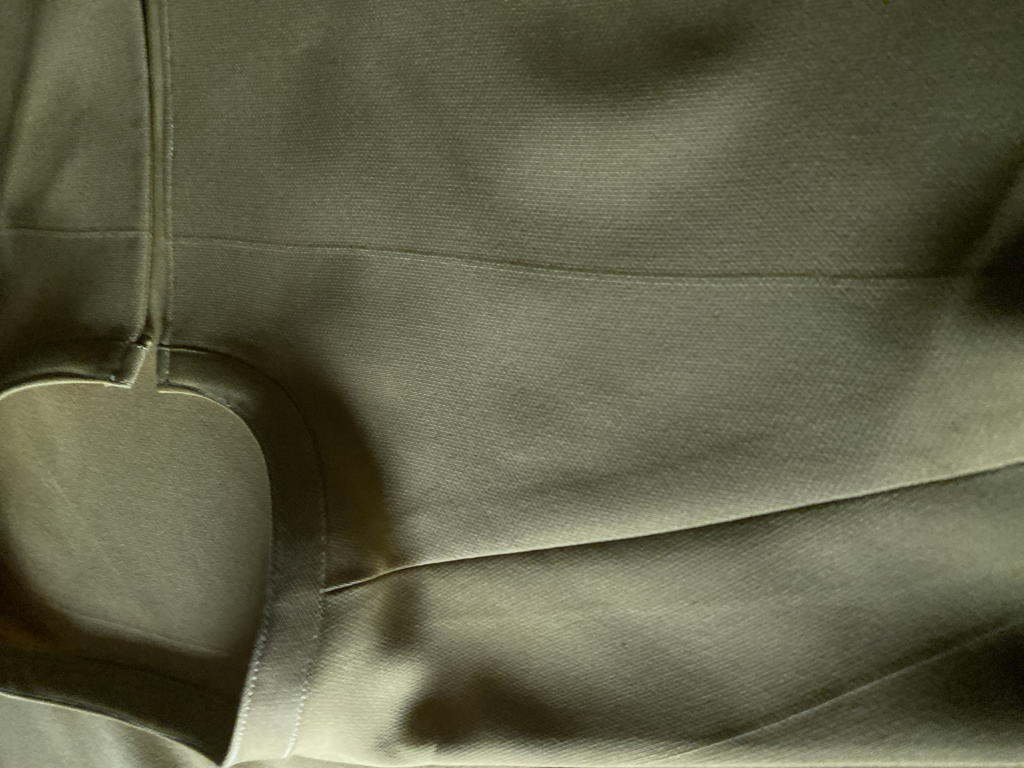 The image shows a close-up view of olive-green fabric, likely from a garment. The fabric has a textured weave and appears to be creased in several places, suggesting it might be part of trousers or a similar item of clothing. A curved seam is visible in the upper left of the image, possibly indicating a pocket opening or a waistband. The lighting creates shadows that highlight the texture and folds of the fabric. There is no discernible urban environment or geographical markers that would indicate a specific location in Jalingo, Nigeria, or any other city.