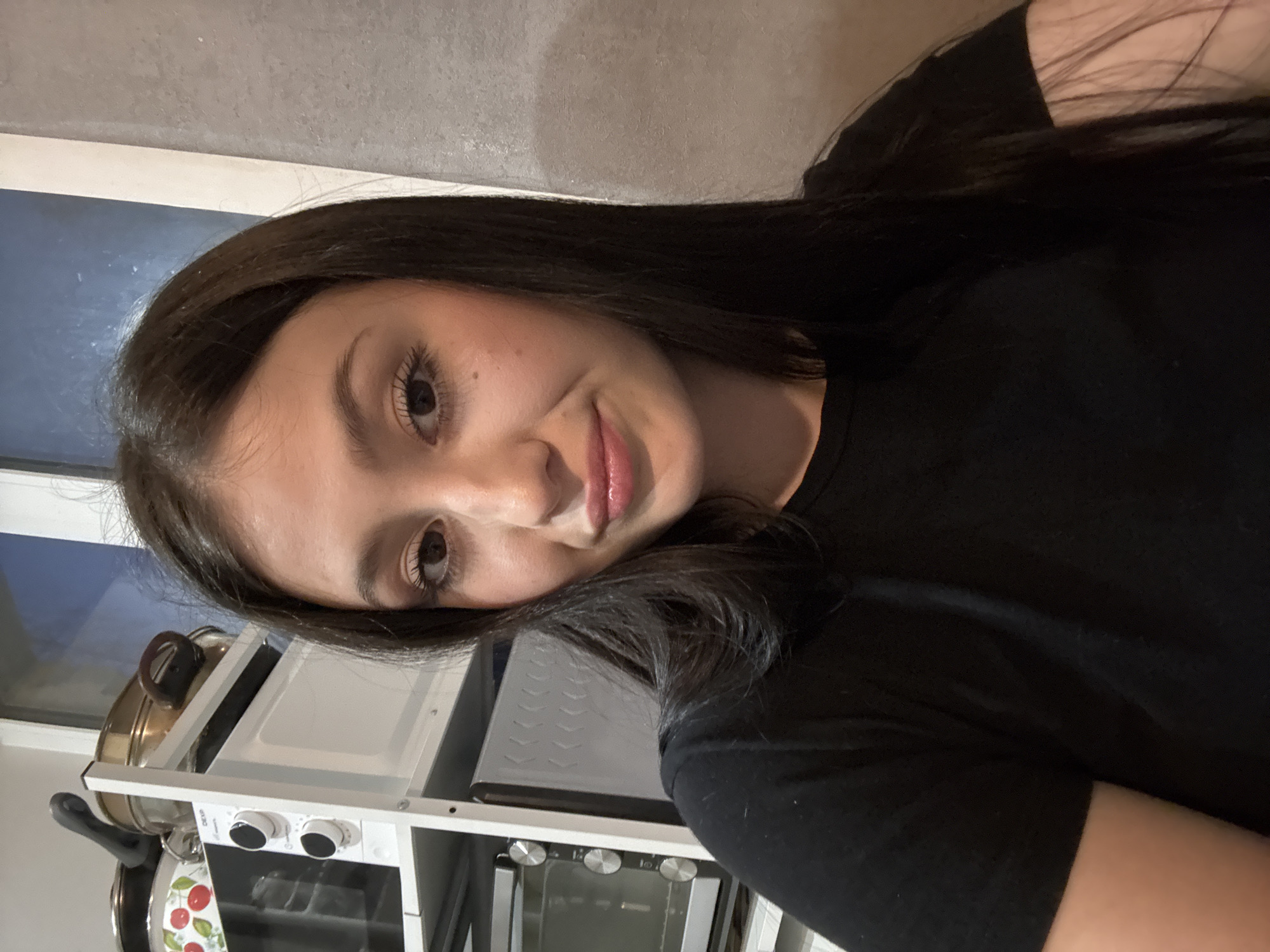 A young woman with long, dark hair is the focal point of the image. She is fair-skinned with light brown eyes and full lips. She is wearing a black t-shirt. The background features kitchen appliances, including a white shelving unit with a toaster oven and other kitchenware. The scene is lit by a combination of natural light from a window and artificial light. There is no definitive information in the image that establishes the location as the city of Chaikovsky, Russia.