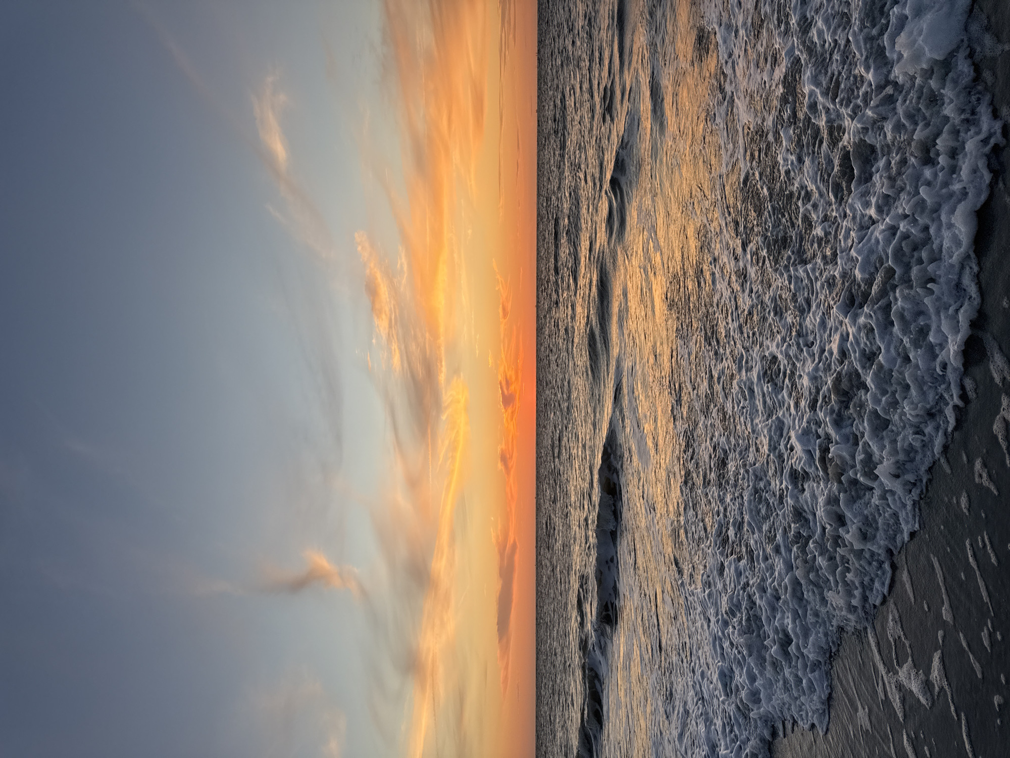 A scenic view of a sunset over the ocean at Langeoog, Germany. The sky is painted with hues of orange, pink, and blue, with wispy clouds scattered across the expanse. The sun, a vibrant orange orb, is setting on the horizon, casting a warm glow on the water. Gentle waves are rolling towards the shore, creating a foamy white surf that laps at the dark sand. The sea reflects the colors of the sky, creating a shimmering, ethereal effect. There are no people or man-made objects visible in the scene, emphasizing the natural beauty of the coastal landscape. The overall impression is one of tranquility and serenity.