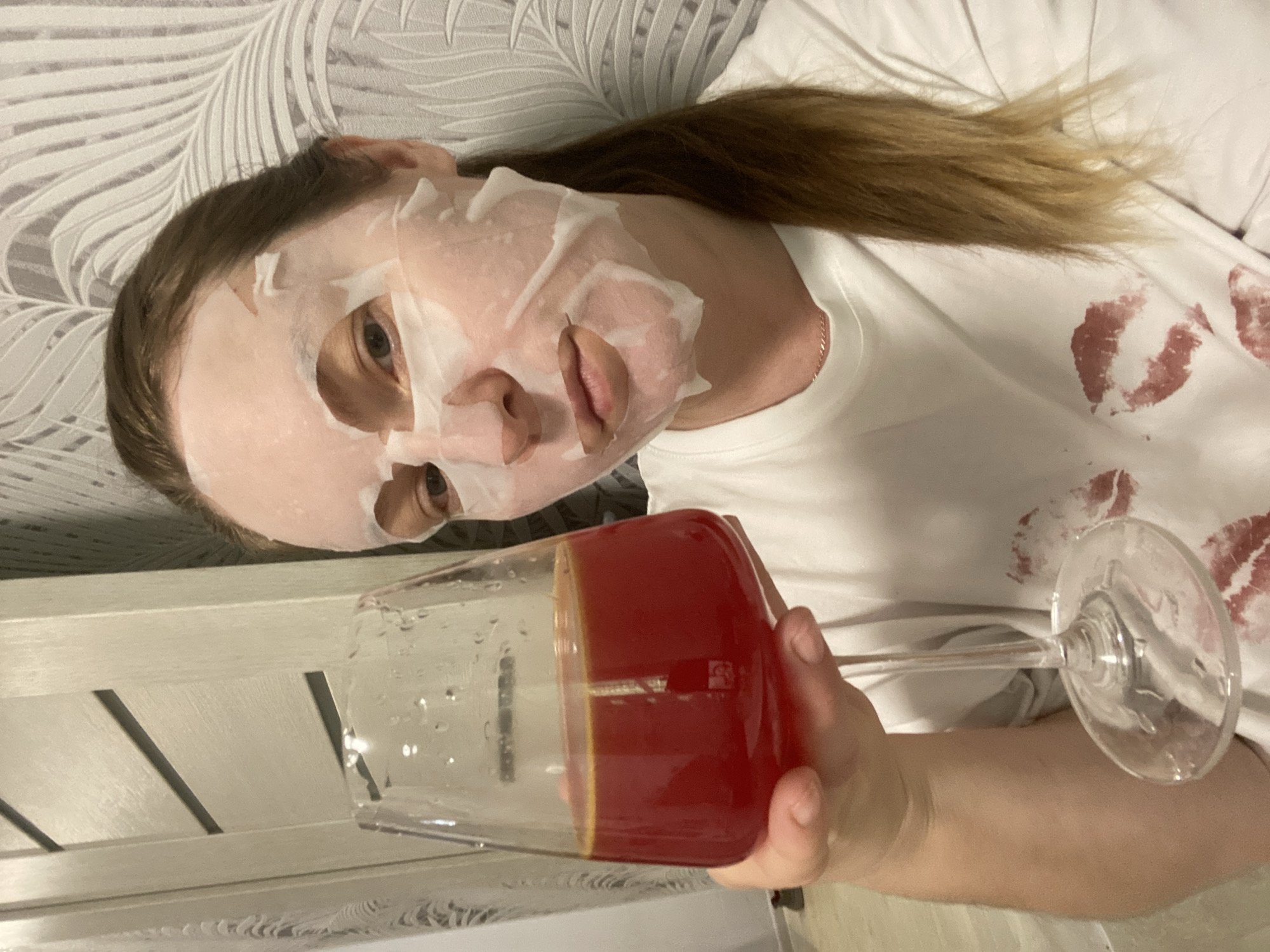A woman with shoulder-length, light brown hair lies supine, wearing a white facial mask and a white t-shirt with red lipstick kisses on it. She is holding a large wine glass filled with a dark red liquid. She appears to be indoors, resting against a surface with a leaf-like pattern and a wooden-slatted structure. There is no evidence indicating a location in Noviy, Russia.