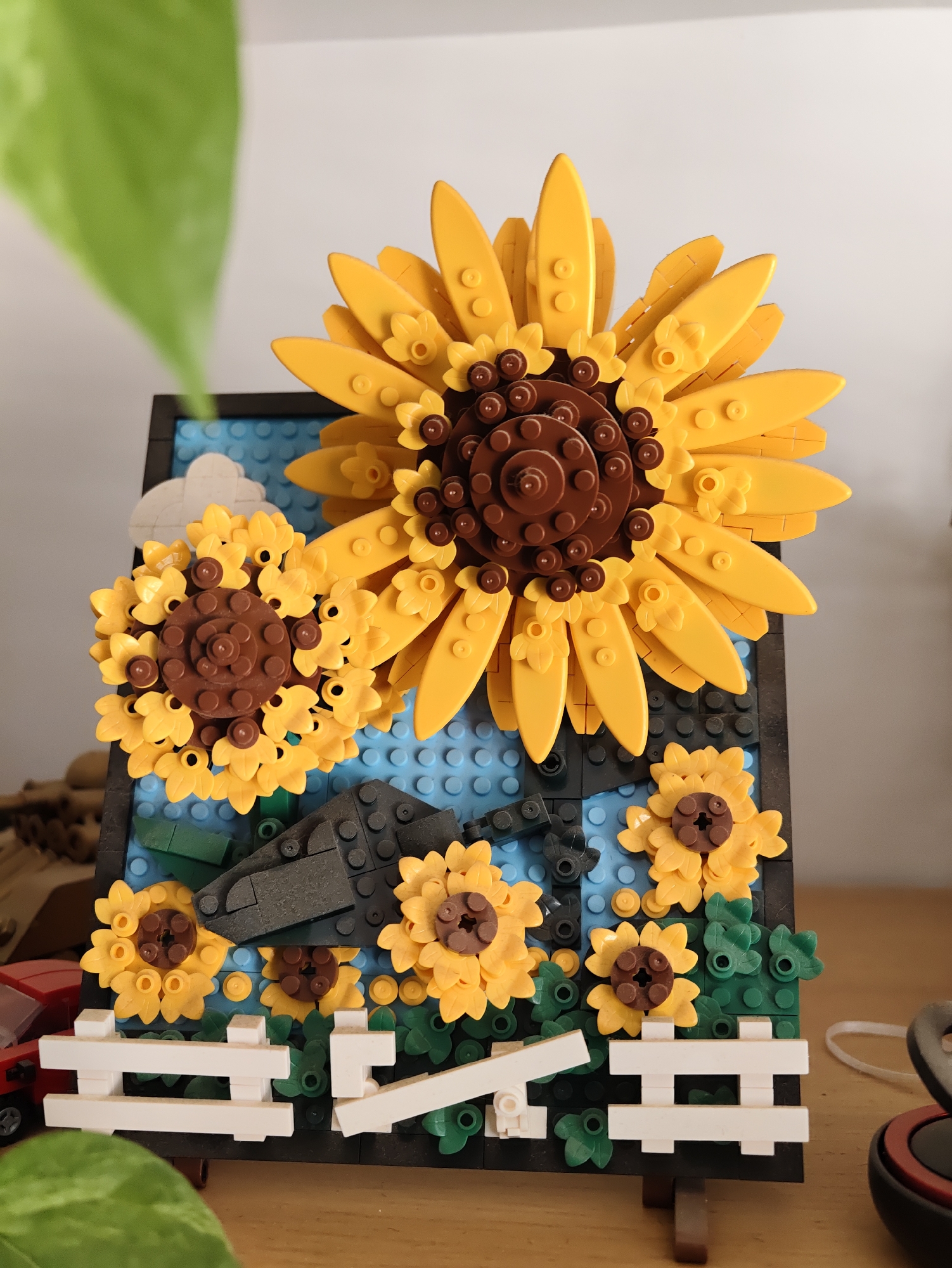 This is a close-up, eye-level, indoor photograph of a LEGO model of sunflowers arranged in a framed display. The main subject is a large sunflower at the top, with bright yellow petals and a dark brown center composed of smaller circular LEGO pieces. Below it, to the left, is a smaller sunflower, also with yellow petals and a brown center. Several other smaller sunflowers are scattered across the lower half of the display, interspersed with green LEGO leaves and blue LEGO pieces representing the sky. A white LEGO fence is visible at the bottom of the display. The LEGO model is propped up on a dark brown stand. The background is a plain white wall, with a large green leaf of a houseplant visible in the upper left corner, out of focus. The lighting suggests an indoor setting, possibly a home or a display area. The scene is static and does not depict any people or actions. The photograph appears to be taken with a smartphone. The image is in focus and well-lit. There is no visible indication of the city of Petah Tikva, Israel, within the image itself.