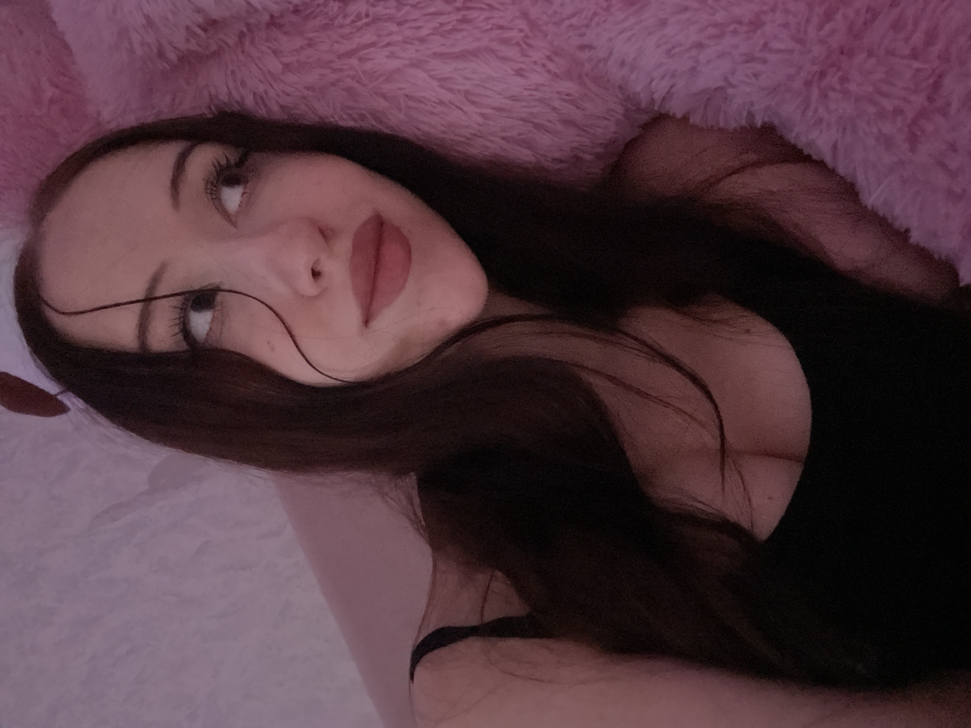 The image shows a close-up, head-and-shoulders shot of a young woman lying down, possibly in bed, with a pink, fluffy blanket or pillow next to her. She has long, dark brown hair, fair skin, and is wearing a black tank top. She is looking upwards and to her right, with her eyes wide open and makeup on her face, including mascara and lipstick. There are what appear to be small, light-colored fuzzy accessories on top of her head, resembling ears. The lighting in the image is soft and pinkish, creating a warm and intimate atmosphere. The background to her left is a lighter pink, textured surface, possibly a wall or bedding. The image does not contain any information or visual cues that would allow for identification of the specific location within Votkinsk, Russia, or any other city.