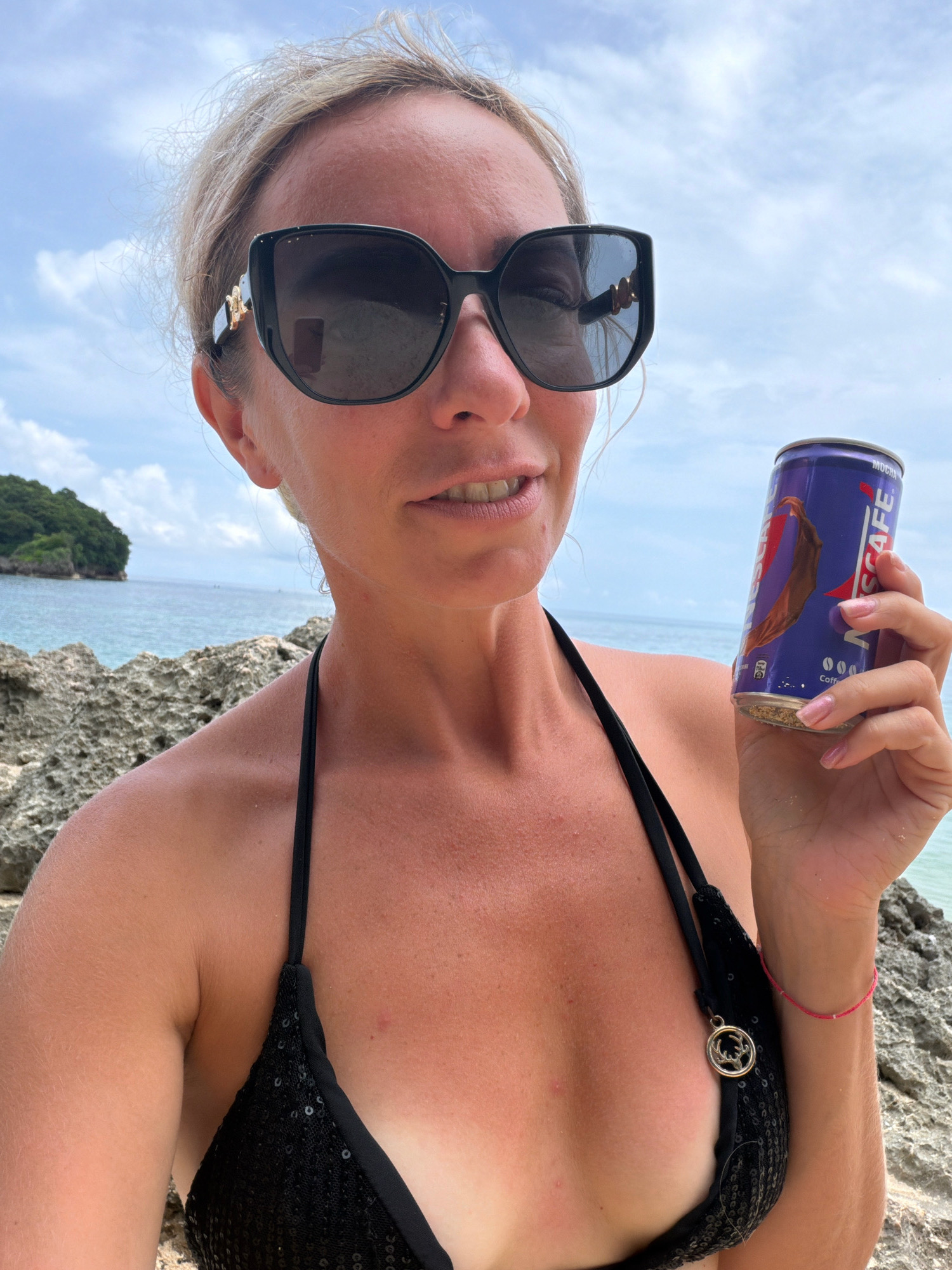 A Caucasian woman is posing for a selfie on a beach in Malay, Philippines. She is wearing a black sequined bikini and oversized sunglasses, with her blonde hair pulled back. In her right hand, she holds a purple can of Nescafé. The background shows a rocky shoreline, the turquoise ocean, and a lush green island in the distance under a cloudy sky.