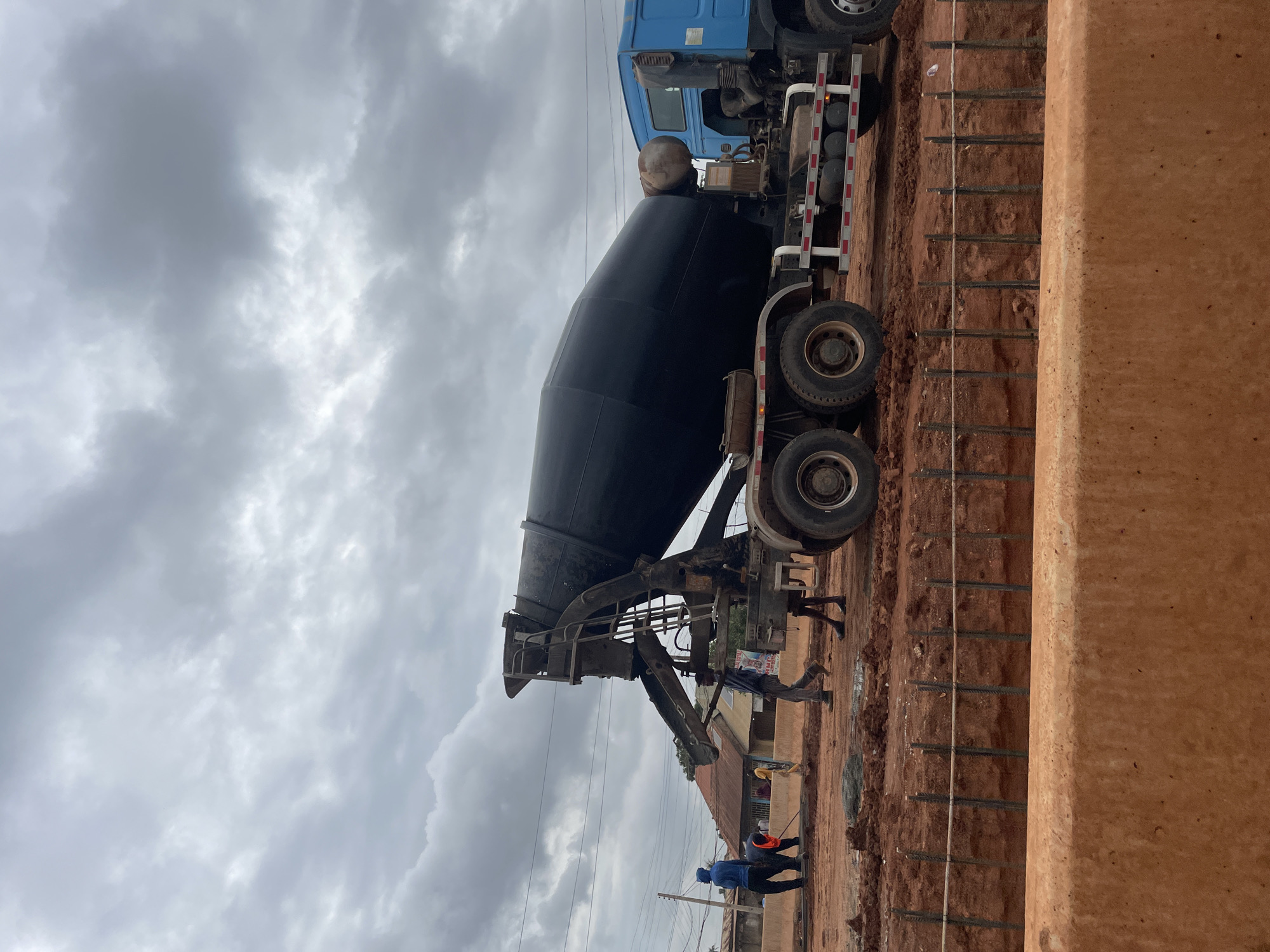 The image depicts a construction site in Minna, Nigeria, under a cloudy sky. A large blue cement mixer truck is parked on red earth, its black mixing drum tilted. Several construction workers are present on the site. Two workers in the foreground appear to be engaged in work on the ground, one wearing a blue hoodie and the other a bright orange vest. Another worker, dressed in a light blue shirt and grey pants, walks near the truck. In the background, there are buildings with corrugated roofs, suggesting a semi-urban or residential area. Power lines are visible against the overcast sky. The ground is uneven, with piles of red dirt and some muddy patches. A section of the construction area shows a foundation being prepared, with vertical rebar protruding from the ground and a rough, orange-colored barrier in the foreground. The overall scene is indicative of ongoing road or infrastructure development.