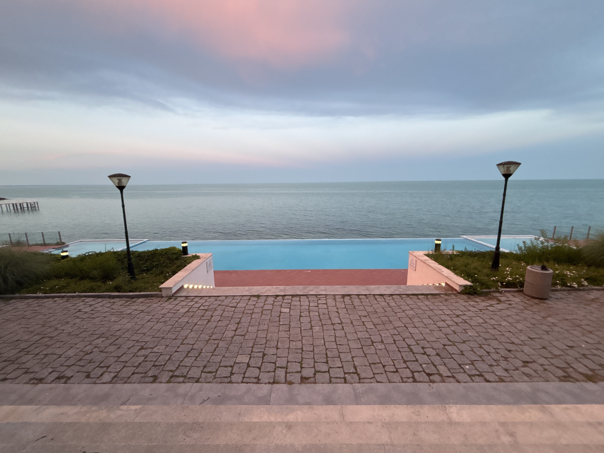 The image depicts an infinity pool overlooking the sea at dusk. The sky is a blend of soft pinks and blues, with scattered clouds. The pool itself is divided into two sections: a lighter blue area in front, and a deeper blue section towards the sea. A red band separates the two sections.
The poolside is paved with cobblestones, and lined with greenery and two lampposts. A concrete trash receptacle is visible to the right. The foreground shows a set of stone stairs leading down to the paved area. A weathered wooden structure, possibly a pier or remnants of one, is visible in the sea to the left. The scene appears to be a tranquil setting, likely a hotel or resort, possibly located along the coast of Varna, Bulgaria, given the architectural style and seaside location. There are no people visible in the image.