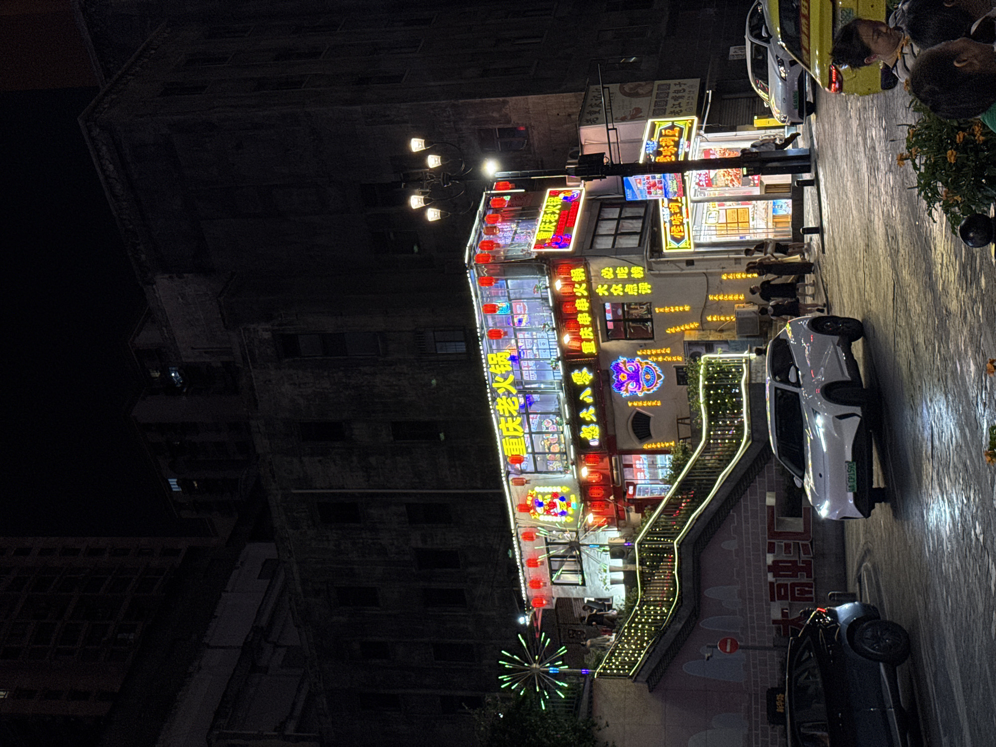 The image shows a busy street scene at night in Yuzhong District, China, with a multi-story building illuminated by numerous neon signs and lanterns. The building appears to be a restaurant or commercial establishment, with text on the signs indicating "Chongqing Old Hot Pot" and other food-related businesses. Several cars are parked or driving on the street in the foreground, and pedestrians are visible walking on the sidewalk and near the entrance of the building. The architecture is a mix of traditional and modern styles, with the older building in the background contrasting with the brightly lit facade of the establishment in the foreground. The street is paved with stone-like material, and the overall atmosphere is lively and vibrant, typical of a commercial district at night.