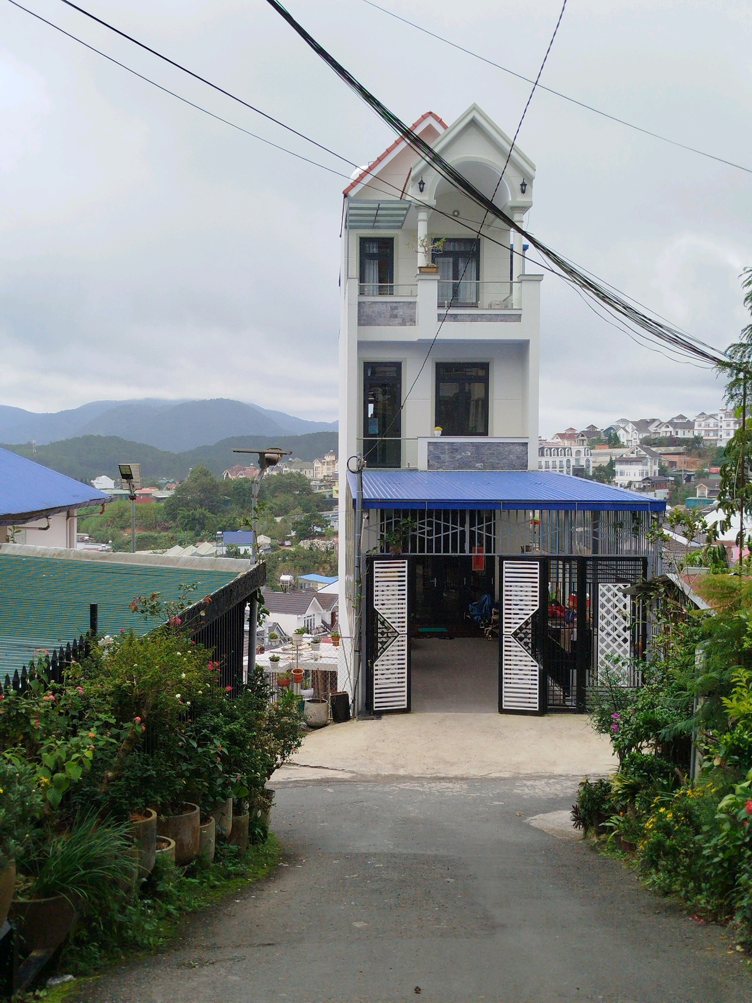 This is a wide-angle, eye-level exterior shot of a narrow, multi-story residential building situated on a hillside in Da Lat, Vietnam. The building is painted white with red-tiled gabled roofs. It features multiple balconies and windows, with prominent utility wires crisscrossing overhead. A blue-roofed awning covers the entrance, which is framed by an ornate black metal gate. In the foreground, a paved pathway leads up to the building, flanked by lush greenery and potted plants. The background reveals a hilly landscape dotted with other residential structures and distant mountains shrouded in mist, characteristic of Da Lat's topography. No people are visible. The sky is overcast, contributing to a diffused light.