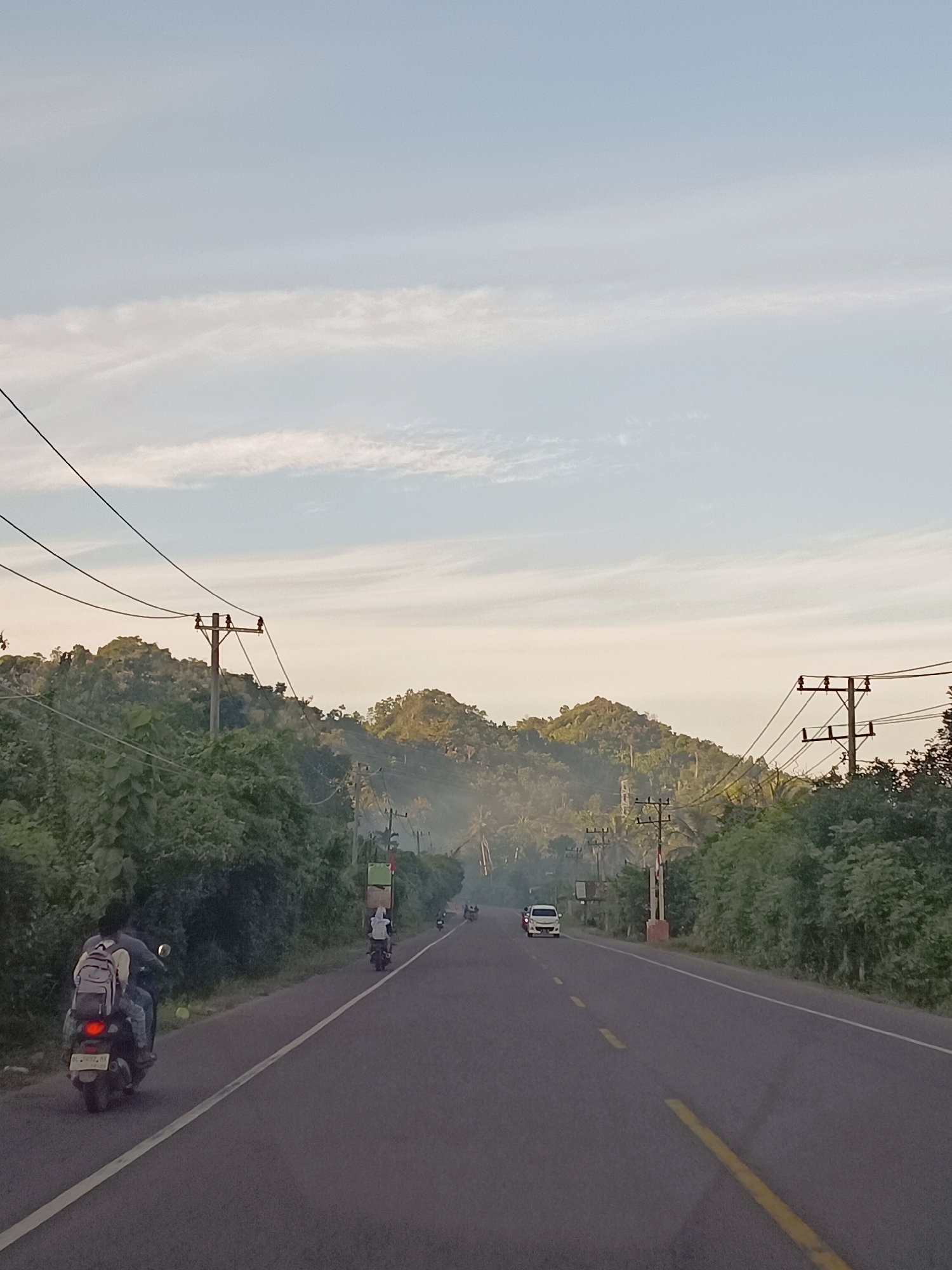 The image captures a scenic view of a road winding through lush green hills under a pale blue sky with wispy clouds. In the foreground, a person rides a scooter on the right side of the road, heading away from the viewer. The scooter has a backpack strapped to its rear. The road is paved with a white line marking the edge and a yellow line dividing the lanes.

Further down the road, other vehicles, including a white car and several more scooters, are visible. Utility poles with wires run along the side of the road, interspersed with dense foliage and trees. The background is dominated by rolling hills covered in vegetation, with a light haze or mist partially obscuring the distant peaks, suggesting a humid or possibly early morning/late afternoon atmosphere. The overall scene depicts a rural or semi-rural setting in Tu, Indonesia, with active transportation and natural landscapes.