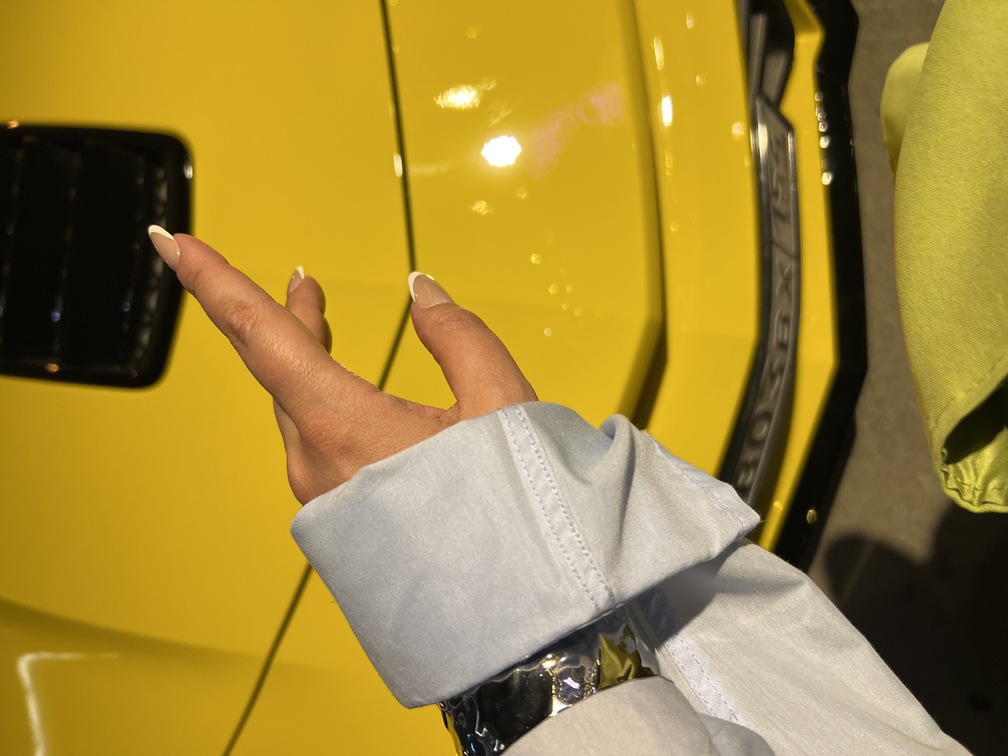 A hand wearing a light blue shirt sleeve and a silver bracelet is extended toward a yellow vehicle. The vehicle has black accents and lettering that includes "ZLX/ZL1." Part of a lime green garment is visible on the right side of the image. The location is not identifiable.