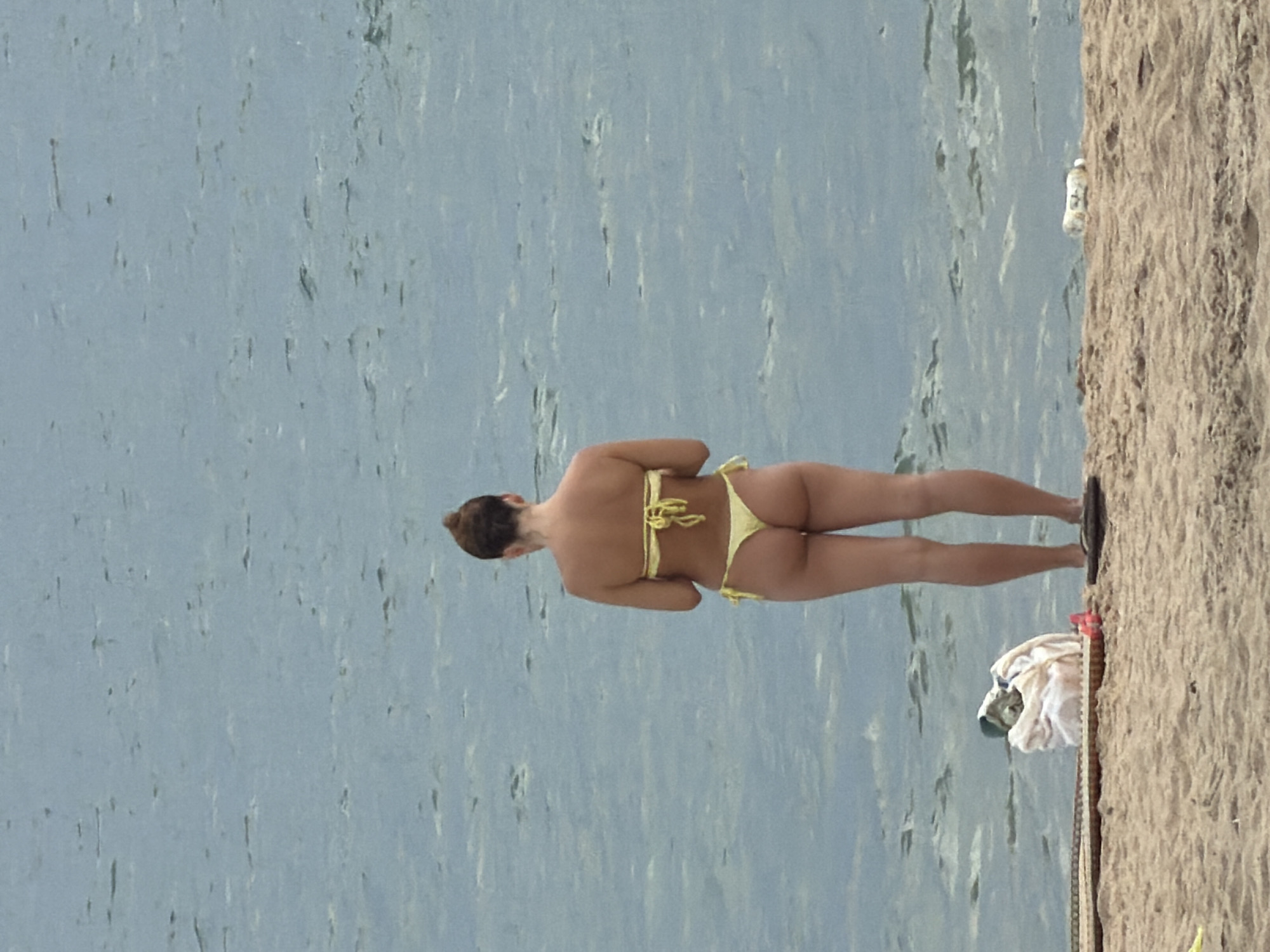 A woman stands on a sandy beach, facing away from the camera, with her back to the ocean. She is wearing a yellow bikini. Her hair is pulled up in a bun. In front of her on the sand is a pile of clothes, possibly a towel and a bag. There are also a couple of flip-flops on the sand. The ocean is calm and blue. The scene appears to be in Nha Trang, Vietnam, a coastal city known for its beaches.