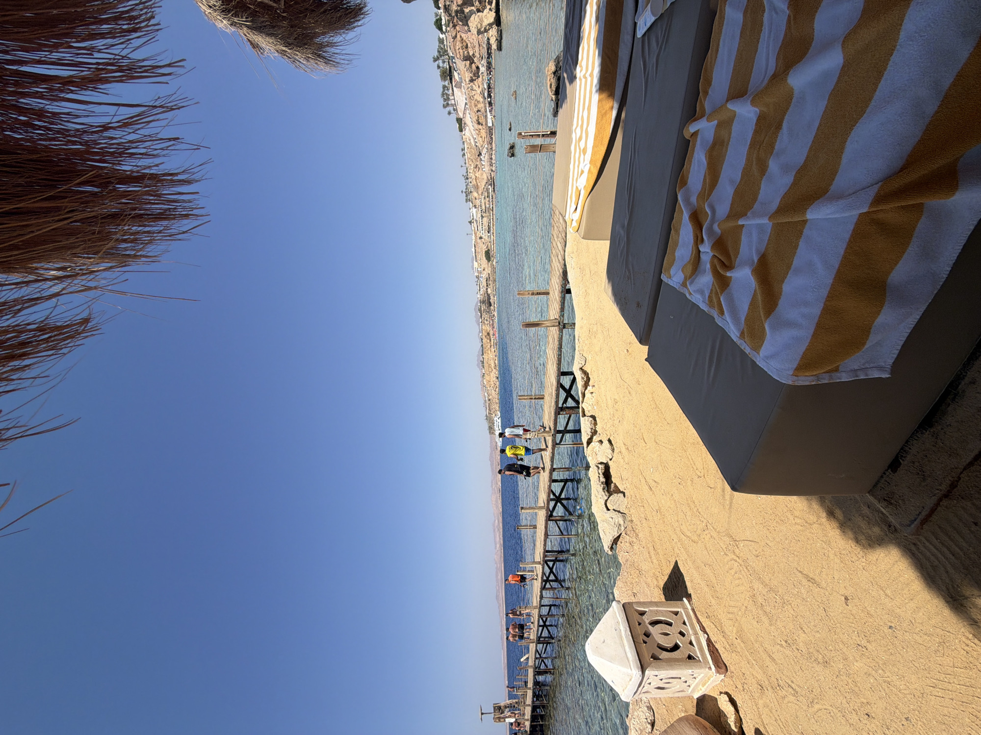 The image depicts a scene at a beach resort in Sharm El Sheikh, Egypt, on a clear, sunny day. In the foreground, two empty lounge chairs with striped towels are set up on the sand, suggesting a relaxed atmosphere. Beyond the chairs, a wooden pier extends into the clear, turquoise water, with several people walking along it. The pier leads to a beach area with rocky outcrops and buildings nestled into the hillside in the background, characteristic of the resort landscape in Sharm El Sheikh. The sky is a vibrant, cloudless blue.