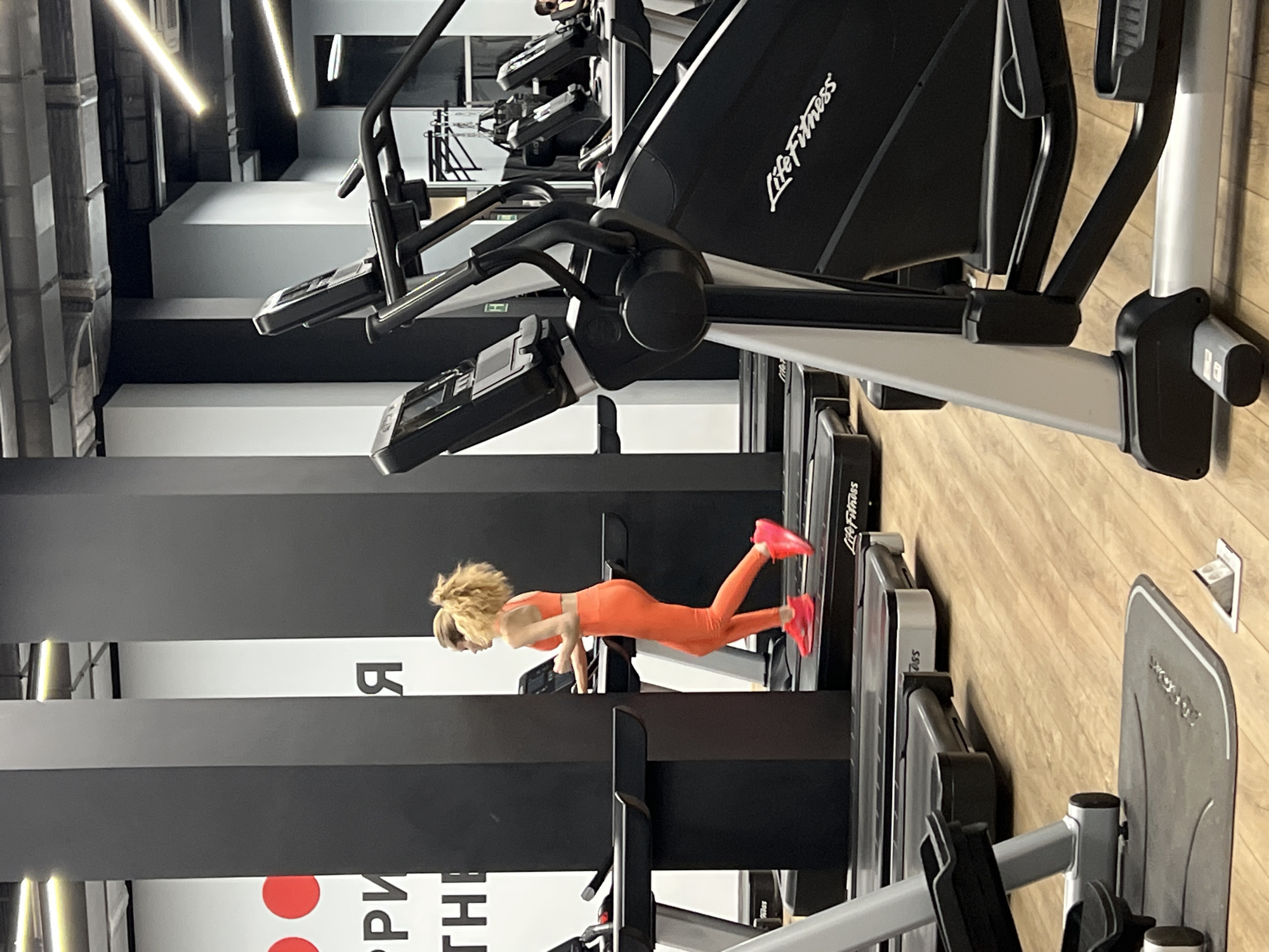 A woman in an orange workout outfit and pink sneakers is doing push-ups on a treadmill in a gym. The gym contains rows of treadmills and elliptical machines. The location may be Moscow, Russia, due to the Cyrillic text on the wall. The gym has bright overhead lighting, black and white wall stripes, and a wood-look floor.