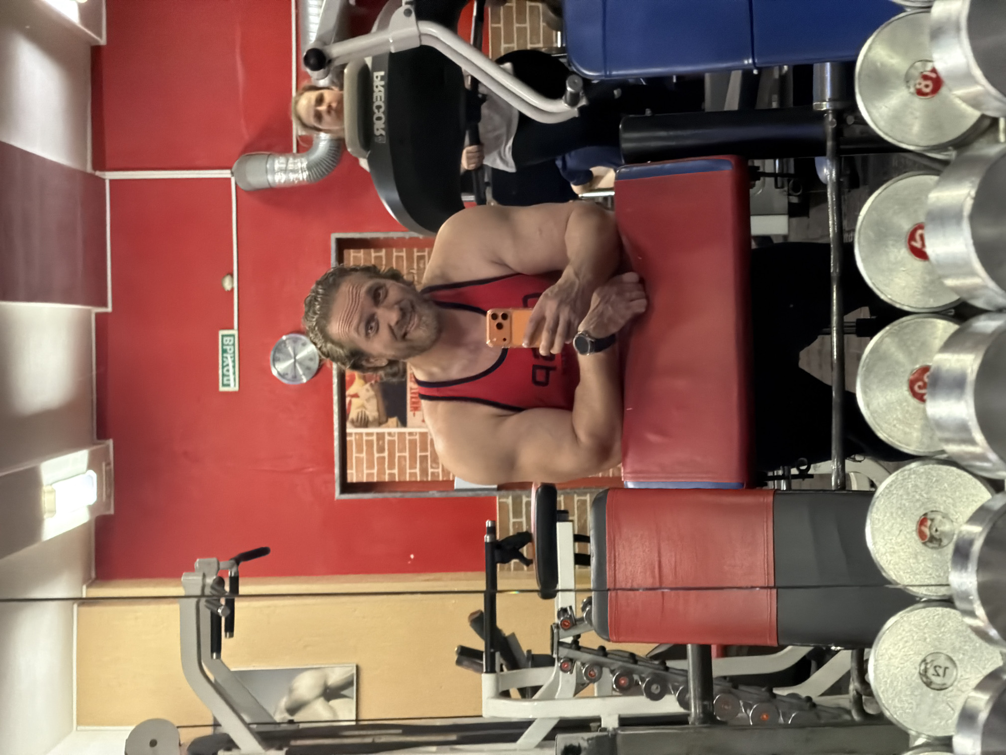 This image depicts a male individual taking a selfie in a gym located in Saint Petersburg, Russia, with the scene reflected in a large mirror.
The primary subject is a muscular adult male, mid-shot, with wavy brown hair and a beard, wearing a red tank top with a black stripe and dark athletic pants. He is smiling and looking directly at the camera, holding an orange smartphone in his right hand to capture the image. His left forearm, adorned with a dark wristwatch, rests on a red and black upholstered pad of a gym machine.
In the background, positioned to the right of the male subject (viewer's left), a female individual is visible, wearing a light-colored top and dark pants. She is operating a large black fitness machine, identifiable as a "PRECOR" brand by its reversed text on the console. Her expression is neutral.
The environment features red-painted walls with sections of lighter, possibly wooden, paneling. A green "ВЫХОД" (EXIT) sign, visible in reverse, is affixed to the red wall, confirming a Russian-speaking location. A silver circular wall clock and a flexible metallic ventilation duct are also visible on the red wall. The ceiling consists of white and red checkered panels with fluorescent tube lighting. Various pieces of gym equipment are present throughout the reflected space, including a dumbbell rack in the foreground with chrome dumbbells displaying numbers such as "5", "8", and "15". A doorway with a brick-patterned frame leads to another area of the gym.