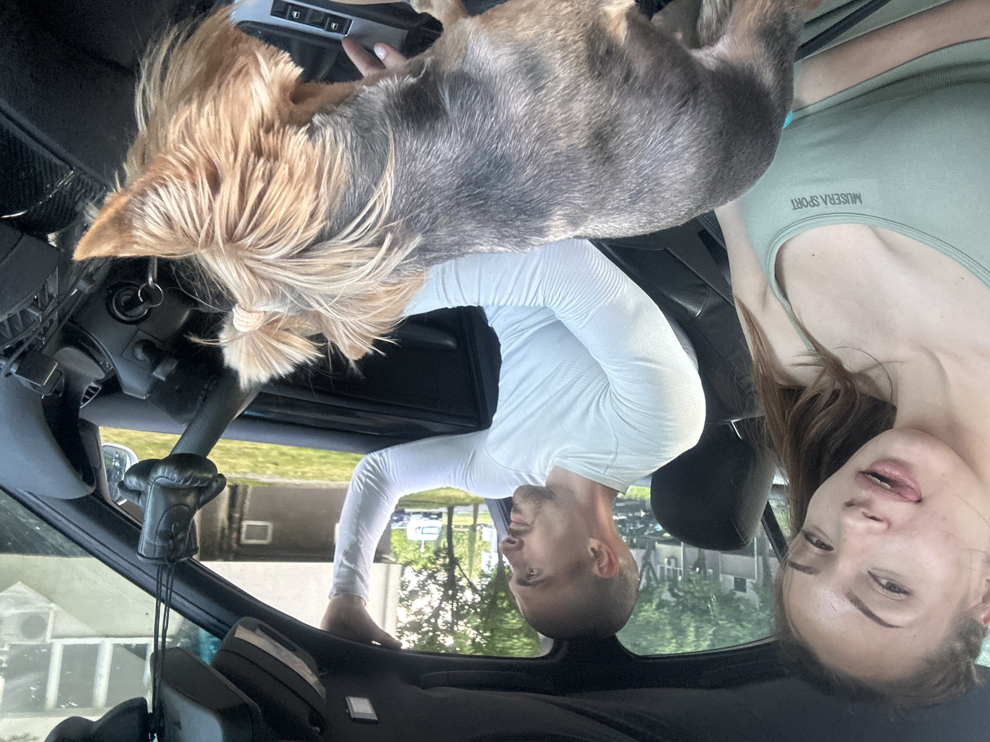 A woman wearing a light green tank top, a man wearing a white long-sleeved shirt, and a small dog are in a car. The woman is in the passenger seat and is looking towards the camera. The man is in the back seat. The dog is on the dashboard of the car. The interior of the car is black. A pair of small boxing gloves is hanging from the rearview mirror. The scene appears to be inside a car in an outdoor location, with a view of buildings and trees through the car windows. Without more detailed landmarks, it is not possible to pinpoint the location within Burgas, Bulgaria.