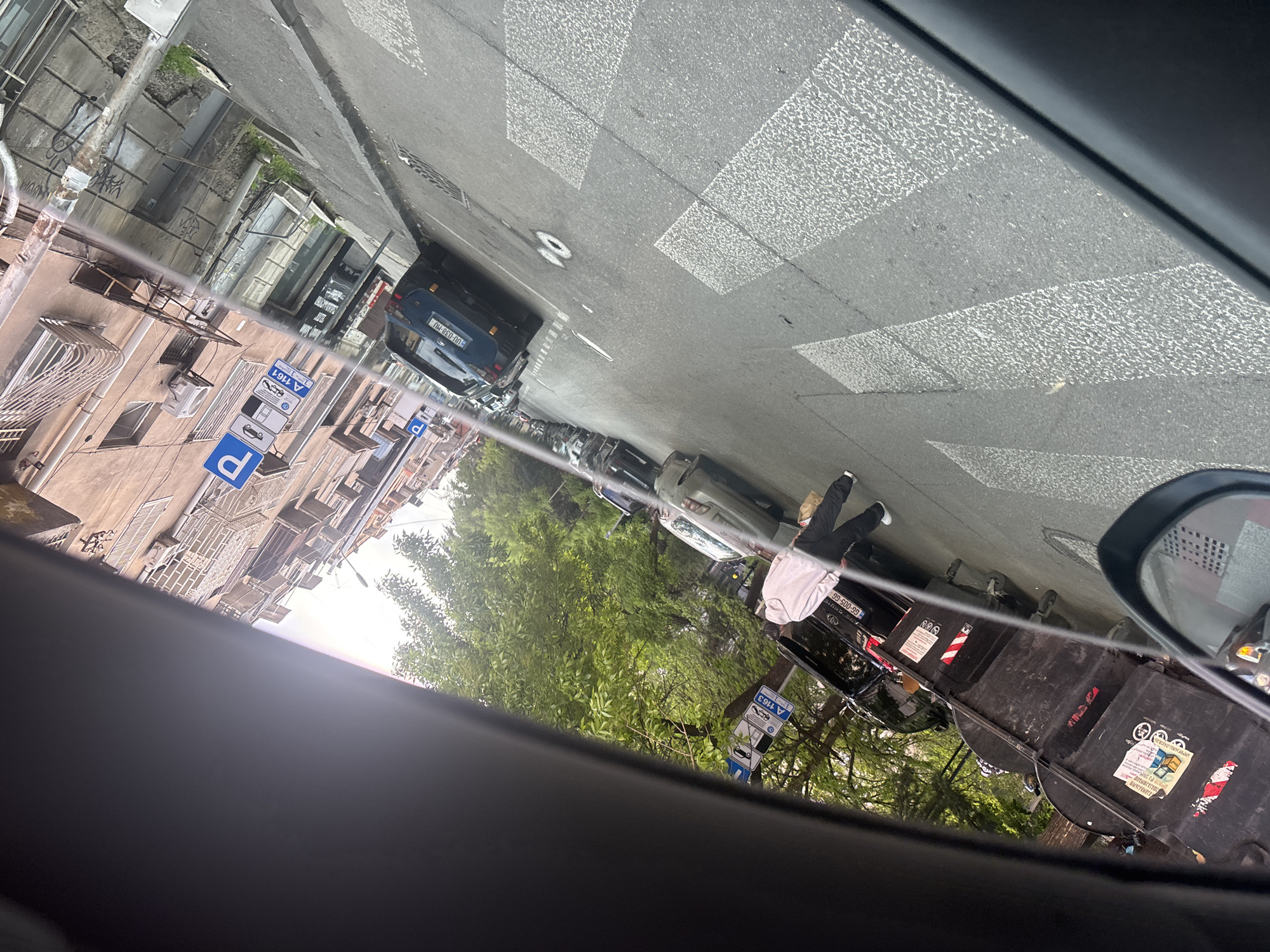 The image, taken from within a vehicle, depicts a street scene in a city, observed through the car window and reflection in a side mirror. The perspective is rotated 90 degrees clockwise from typical orientation.
On the left side of the street (if rotated correctly), multi-story buildings with balconies and air conditioning units line the pavement. Several cars are parked along this side. One notable parking sign is blue with a white 'P' and features a white rectangular sign below it displaying "A 1161" alongside icons for different vehicle types (car, motorcycle).
The center of the image shows the asphalt road with white crosswalk markings. On the right side of the street, more cars are parked alongside the curb. A person, whose lower body (legs in dark pants, white sneakers) and upper torso (light-colored shirt/jacket) are visible, stands between a black car with license plate "GG-005-GG" and a series of large, black trash bins. One of the bins has a white sticker with Georgian text and a logo, and another appears to have a label with "БИЧСРВЫЕСЕ", possibly Cyrillic or distorted Georgian. Another parking sign, similar to the one on the opposite side, displays a blue 'P' and "A 1163". The rear of a blue car with license plate "UU-038-MU" is also visible further down the street. Green trees line parts of the street, suggesting some greenery in the urban environment.
The weather appears to be daylight, possibly overcast, with ample natural light. The architecture and visible text, particularly the Georgian license plates and script on signs, indicate the location is Tbilisi, Georgia. The overall activity is a typical urban street scene with parked vehicles and a pedestrian.