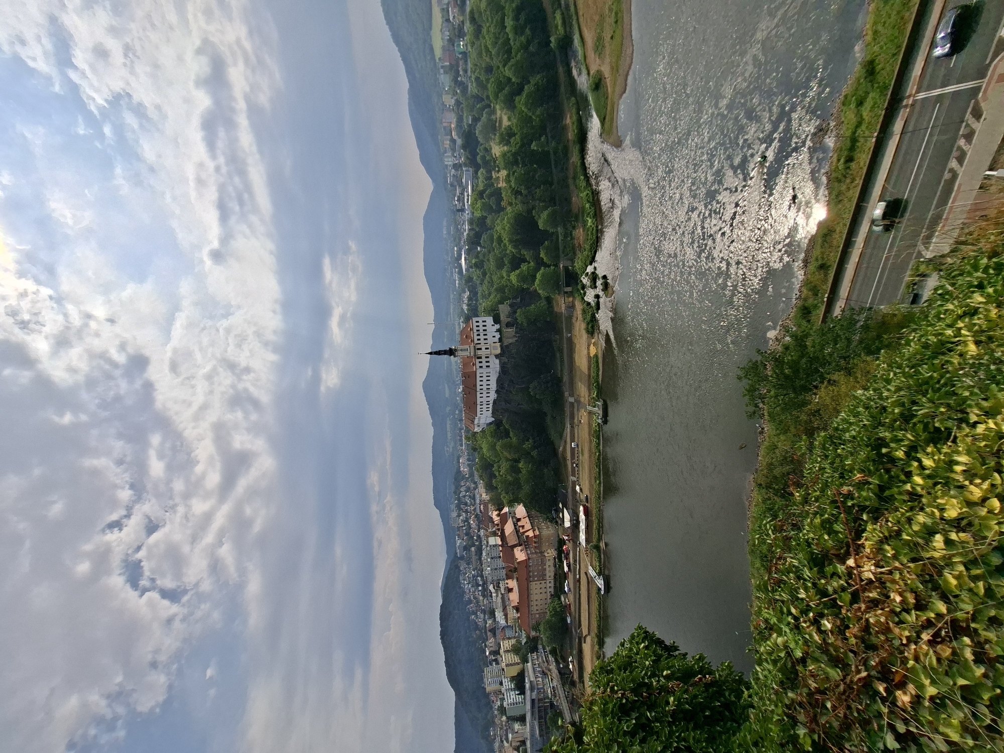 The image presents a wide-angle, elevated view of the city of Děčín, Czechia, with a focus on the Elbe River and a prominent castle complex. In the foreground, lush green vegetation, predominantly ivy, covers a steep embankment. Below this, the Elbe River flows from left to right, its surface reflecting the sunlight breaking through scattered clouds.
Along the riverbank opposite the vegetation, a road runs parallel to the water, with two cars visible. Beyond the road, the city of Děčín unfolds. Several multi-story buildings with red-tiled roofs are clustered together, some appearing older and more traditional, while others are modern concrete structures. A bridge spans a section of the river further into the cityscape.
Dominating the middle ground is the Děčín Castle, situated on a rocky outcrop amidst dense green trees. The castle features white walls and a tall, dark-spired tower. Beyond the castle and the immediate city, rolling hills covered in vegetation form the backdrop, their outlines softened by the distance and atmospheric conditions. The sky is partly cloudy, with patches of blue sky visible and sunlight intermittently illuminating the scene, creating bright reflections on the river. The overall impression is of a scenic riverfront city with historical architecture nestled within a natural landscape.