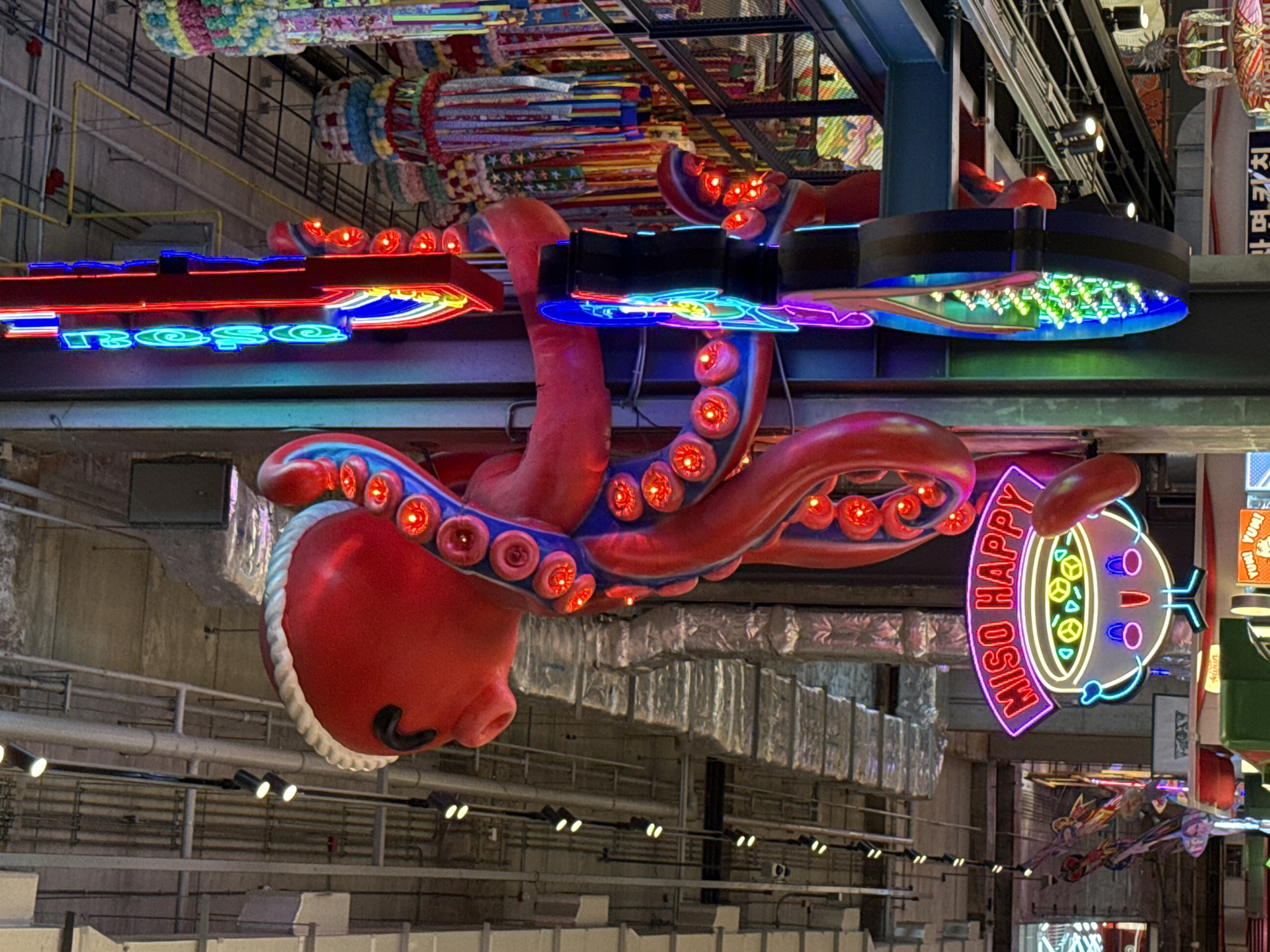 The image shows an upside-down view of decorations at a shopping mall in Bangkok, Thailand. A large, red octopus sculpture with blue accents and glowing red suckers hangs from the ceiling. Above the octopus is a neon sign spelling "roso" in blue and green. To the right of the octopus, a neon sign reads "MISO HAPPY" next to an cartoon bowl of miso soup. In the background, there are various other decorations, including colorful fabric and metal structures, and the architecture of the shopping mall is visible.