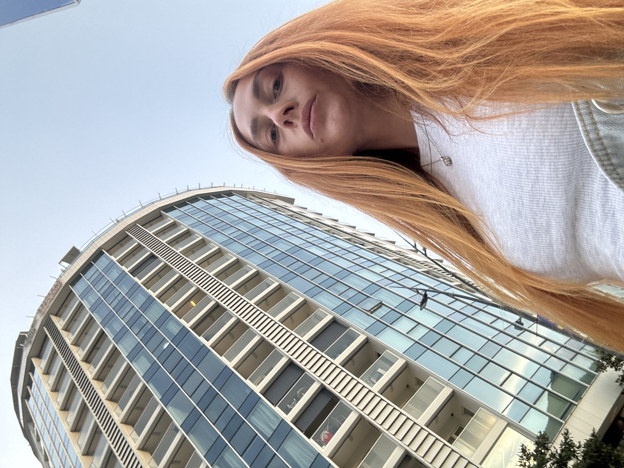 An outdoor, low-angle, medium shot captures a woman with long, ginger hair and a fair complexion. She is wearing a white, ribbed t-shirt and a silver necklace with a small pendant. Her expression is neutral as she looks directly at the camera. Behind her, a modern, multi-story building with a curved facade dominates the frame. The building features numerous balconies and large windows with blue glass, reflecting the sky. The overall environment suggests an urban setting in Budva, Montenegro, under a clear sky.